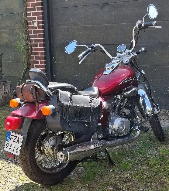 Czerwony motocykl z czarnym siedzeniem i frędzlami, z tablicą rejestracyjną FZA 9AY6. Motocykl posiada dwa lusterka, pomarańczowe kierunkowskazy, chromowane elementy i brązową skórzaną torbę na tylnej części. Znajduje się na trawie i żwirze, na tle ceglanej ściany i ciemnego drewnianego ogrodzenia.