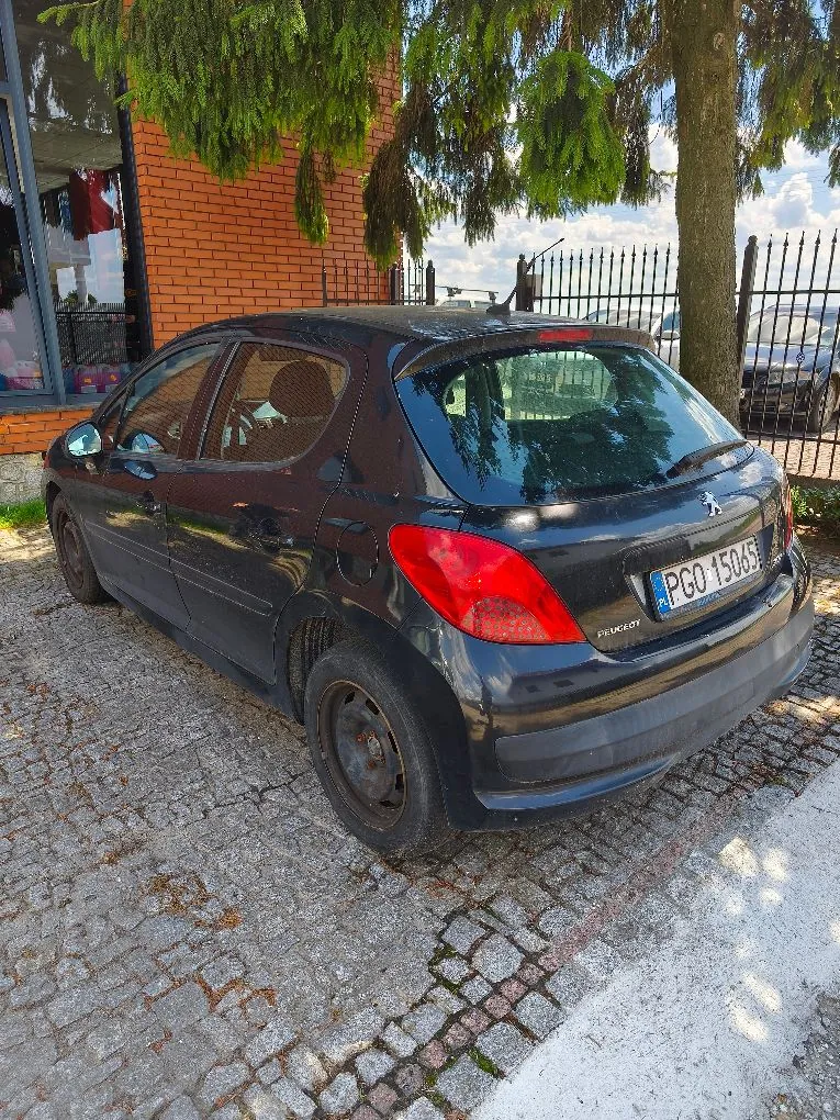 Czarny samochód marki Peugeot 207 zaparkowany tyłem na kostce brukowej. Pojazd znajduje się obok ceglanego budynku i pod drzewem. Na tylnej klapie widoczna jest tablica rejestracyjna o numerze PGO 15065 oraz logo marki Peugeot.