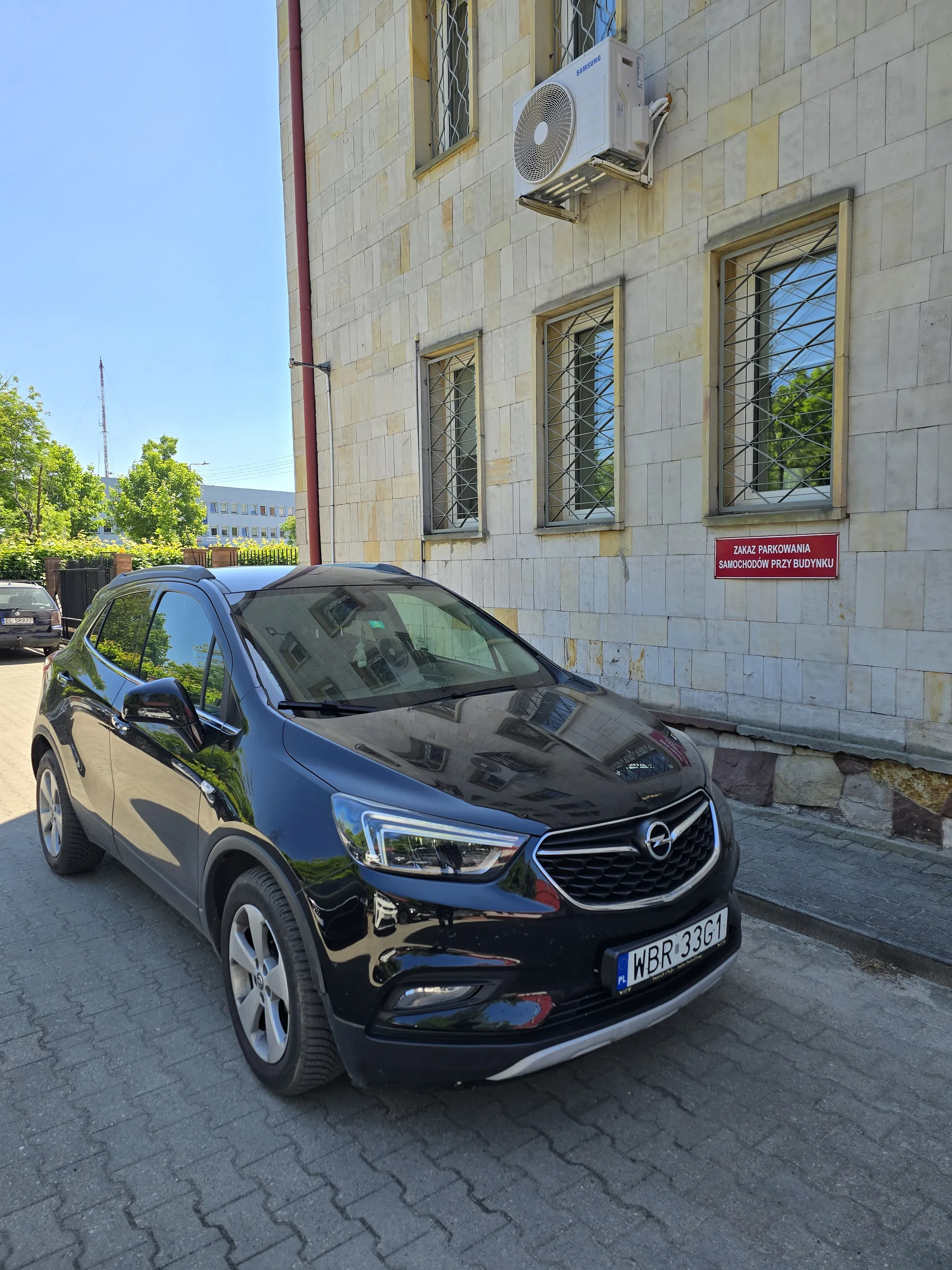 Czarny samochód marki Opel z tablicą rejestracyjną WBR 33G1 zaparkowany na brukowanej powierzchni obok budynku o kamiennej elewacji. Budynek posiada trzy okna z metalowymi kratami, czerwony znak z białym napisem "ZAKAZ PARKOWANIA SAMOCHODÓW PRZY BUDYNKU" oraz zewnętrzny klimatyzator. W tle widoczne są drzewa, niebo oraz fragment innego budynku.