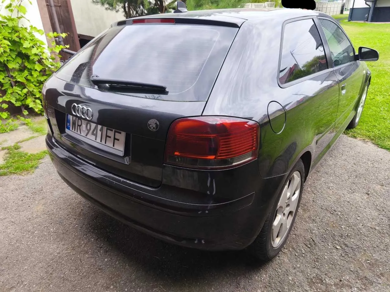 Tylny widok czarnego samochodu marki Audi w nadwoziu hatchback, zaparkowanego na podjeździe. Samochód posiada polską tablicę rejestracyjną o numerze WR 941FF. W tle widoczna jest zieleń oraz fragment budynku.