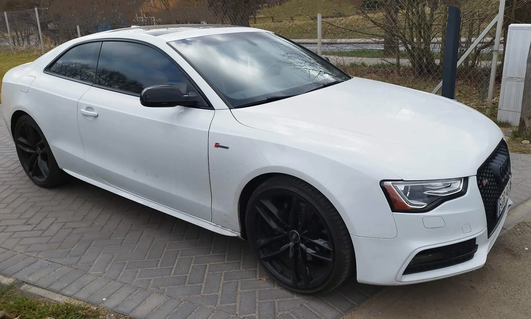 Biały samochód marki Audi w nadwoziu coupe, zaparkowany na kostce brukowej. Pojazd posiada przyciemniane szyby oraz czarne felgi. Na przednim błotniku widoczne jest małe czerwone logo S. W tle znajduje się ogrodzenie z siatki oraz drzewa bez liści.