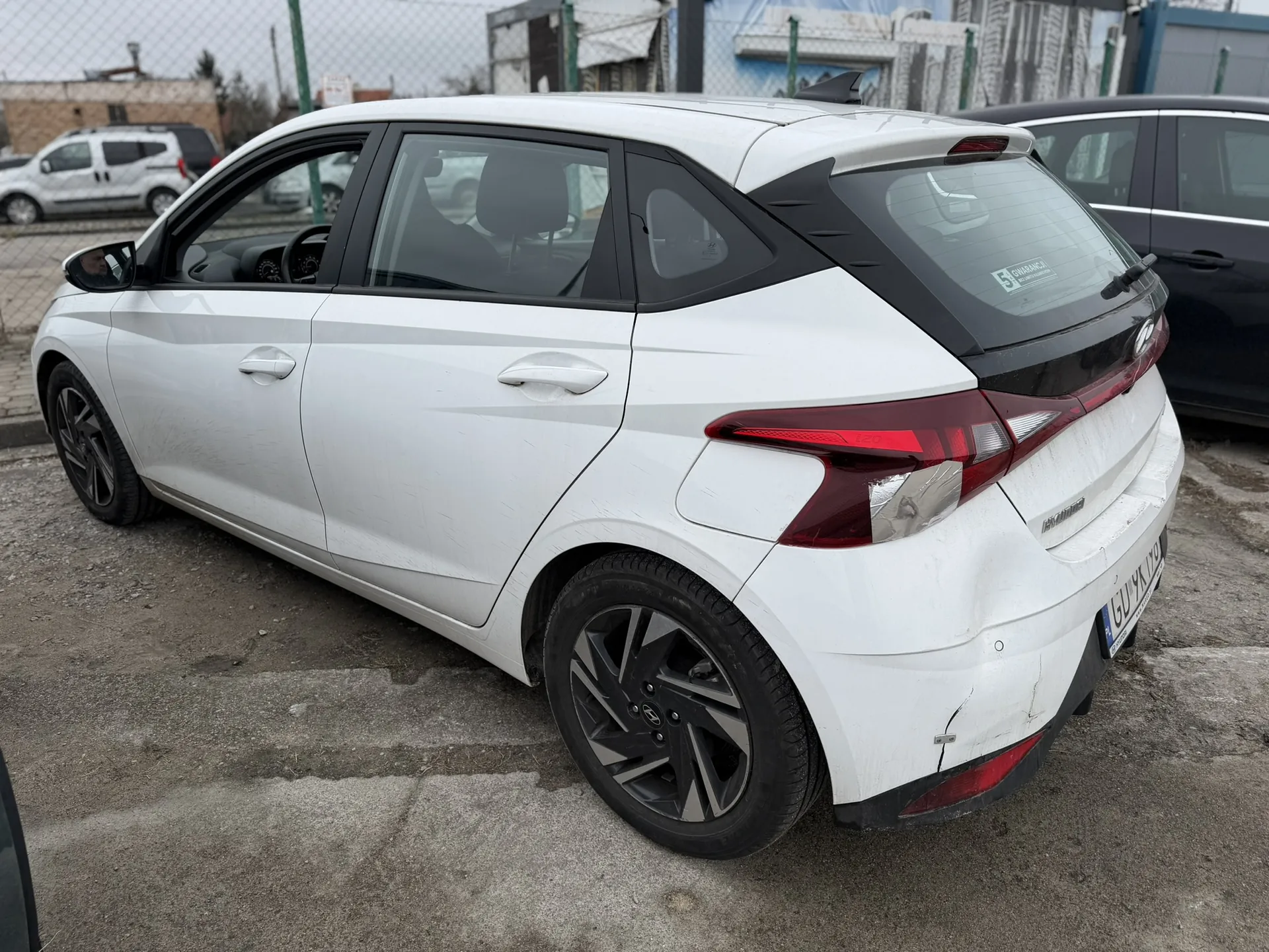 Biały samochód hatchback marki Hyundai, widok z tyłu i boku. Widoczne uszkodzenie na tylnym zderzaku. Czarne akcenty na oknach, czerwone światła tylne. Samochód zaparkowany na betonowej powierzchni. W tle inne pojazdy, ogrodzenie siatkowe oraz budynki. Naklejka z napisem '5 GWARANCJI' na tylnej szybie. Tablica rejestracyjna na tylnej części pojazdu.