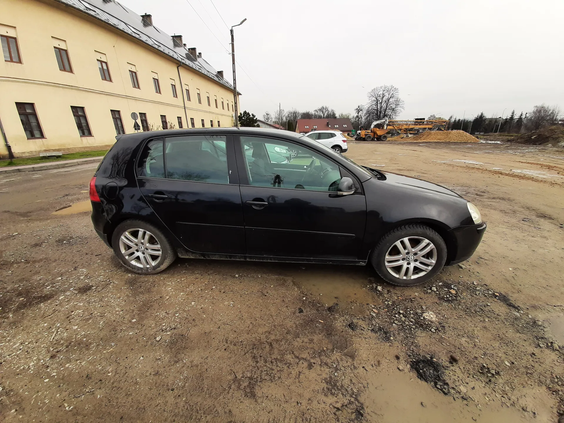 Czarny samochód osobowy typu hatchback marki Volkswagen zaparkowany na błotnistym terenie. W tle żółty budynek z wieloma oknami, biały samochód, maszyny budowlane oraz sterta ziemi. Niebo jest zachmurzone.