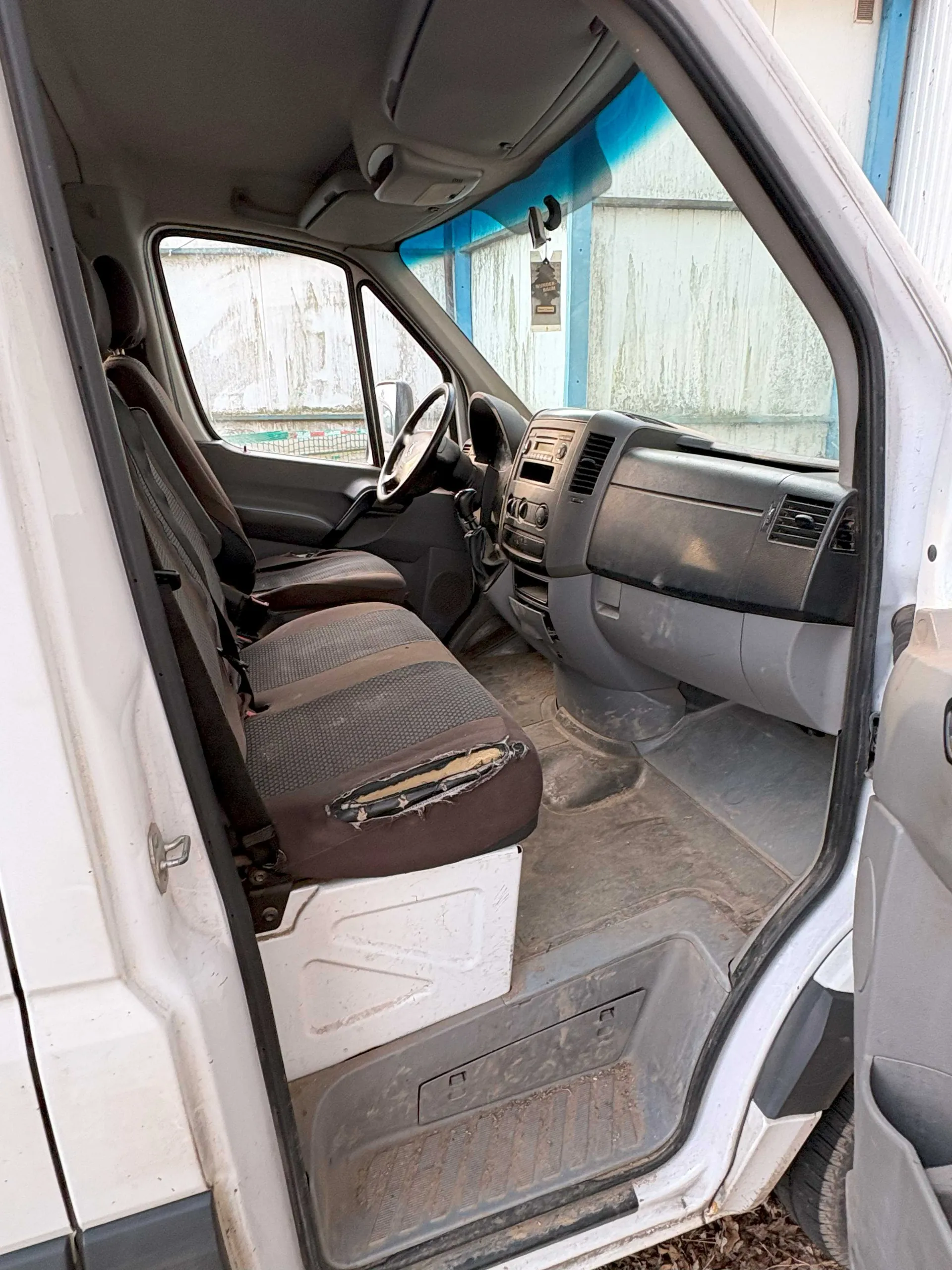 Wnętrze furgonetki z otwartymi drzwiami. Widoczne są przednie siedzenia, z których pasażerskie ma uszkodzoną tapicerkę z odsłoniętym pianką. Kierownica, deska rozdzielcza z przyciskami i radio, oraz brudna podłoga. Zewnętrzna część furgonetki jest biała. W tle budynek z niebieskimi elementami i zniszczoną ścianą.