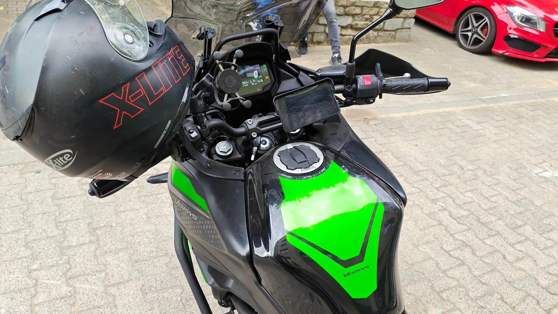 Widok z perspektywy kierowcy na czarny motocykl Kawasaki Versys z zielonymi akcentami na baku. Na kierownicy zamocowany jest czarny kask marki X-lite z czerwonym napisem oraz uchwyt z telefonem. Widoczny jest panel instrumentów motocykla oraz fragment czerwonego samochodu w tle po prawej stronie.