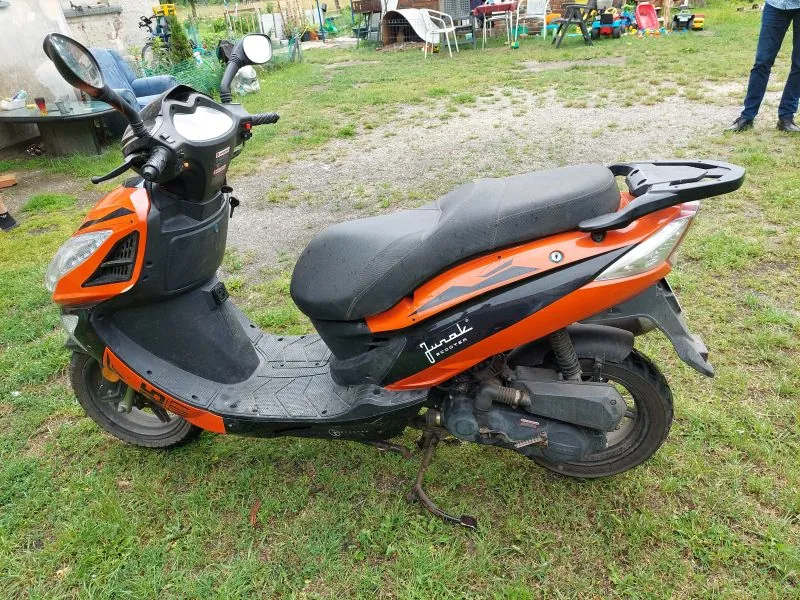 Pomarańczowo-czarny skuter marki Junak Scooter zaparkowany na trawie. Pojazd ma czarną kanapę, przednią tarczę z reflektorem oraz dwa lusterka wsteczne. Na boku skutera widnieje napis 'Junak SCOOTER'. W tle widać ogród z różnymi przedmiotami, takimi jak krzesła i zabawki dziecięce.