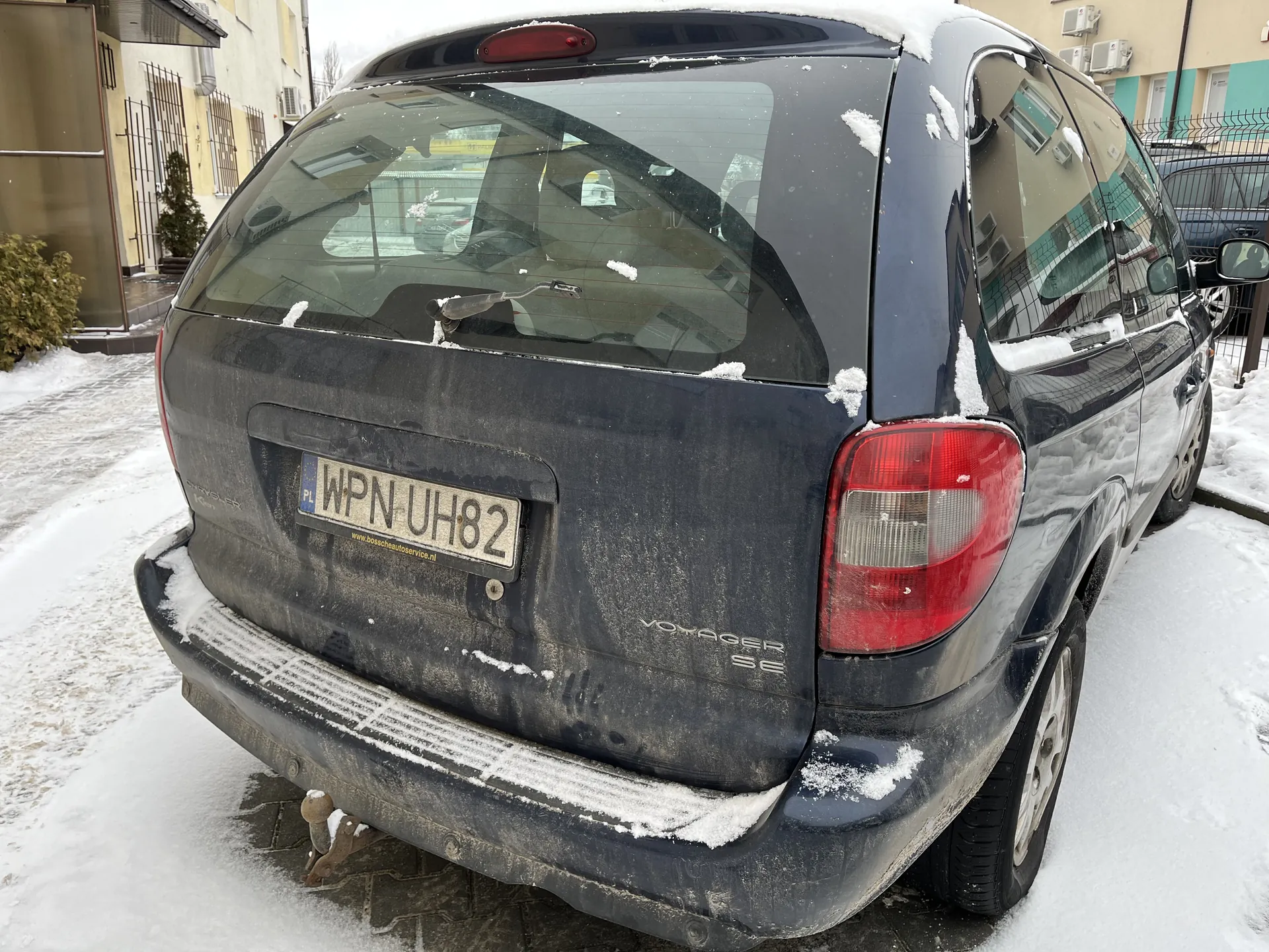 Czarny minivan Voyager SE z tablicą rejestracyjną WPN UH82 i adresem www.boschautoservice.nl. Samochód pokryty śniegiem, zaparkowany na śnieżnej ulicy. W tle budynki i inne pojazdy. Widoczne ślady brudu na karoserii.