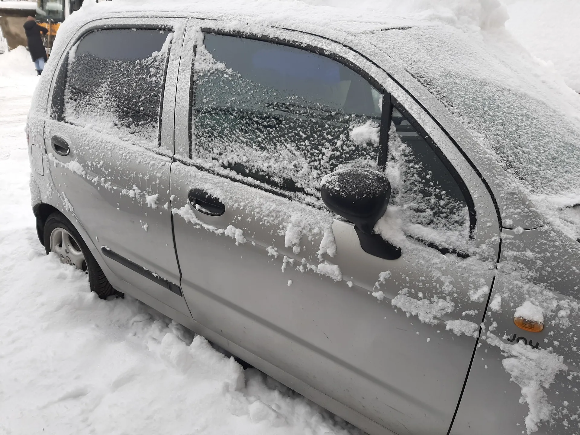 Srebrny samochód pokryty śniegiem, stojący na śnieżnej nawierzchni. Widoczne są zasłane śniegiem szyby, lusterko boczne, klamki drzwi oraz napis 'Joy' na przedniej części pojazdu. W tle osoba w ciemnym płaszczu i niebieskich spodniach oraz budowla.