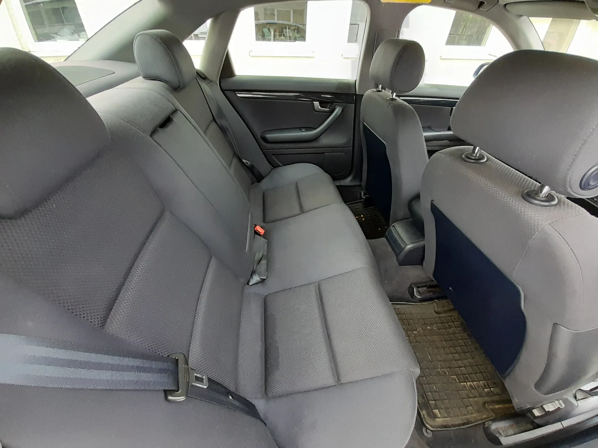 Wnętrze samochodu z widokiem na tylne siedzenia. Szare tkaninowe fotele z zagłówkami, widoczne pasy bezpieczeństwa, w tym jeden z czerwonym zapięciem. Na podłodze czarne gumowe dywaniki. Drzwi samochodu są otwarte, widać fragmenty zewnętrznych ścian budynku przez okna. Wnętrze jest czyste, bez dodatkowych przedmiotów.