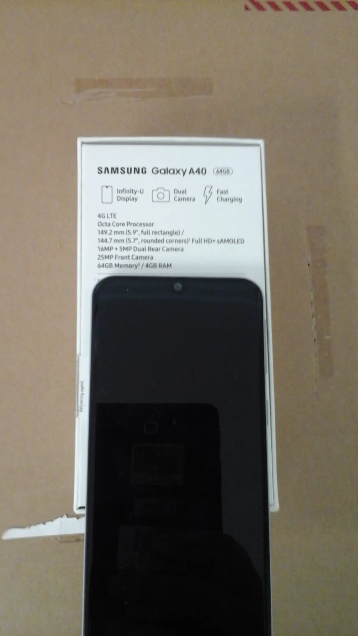 Opakowanie telefonu Samsung Galaxy A40 (64GB) na brązowym tle. Na opakowaniu widoczne informacje: SAMSUNG Galaxy A40, 64GB, Fast Charging, Dual Camera, Infinity-U Display, 4G LTE, Octa Core Processor, 149,2 mm (5,9", full rectangle), 144,7 mm (5,7", rounded corners) Full HD+ sAMOLED, 16MP + 5MP Dual Rear Camera, 25MP Front Camera, 64GB Memory / 4GB RAM. Obok opakowania znajduje się czarny telefon z widocznym ekranem i przyciskiem domowym. W tle brązowa powierzchnia z czerwonymi pionowymi paskami po lewej stronie.