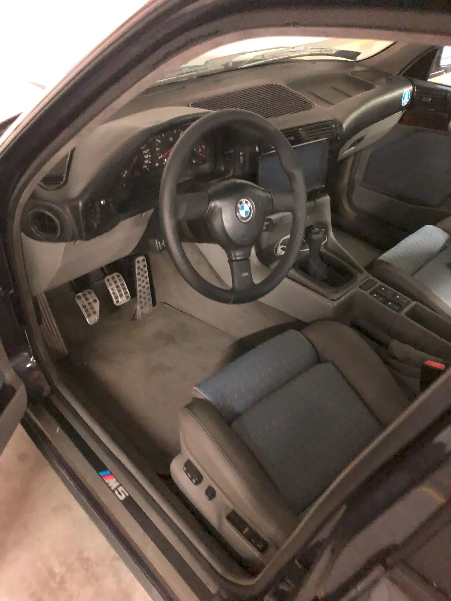 Wnętrze samochodu marki BMW z widokiem na stronę kierowcy. Widoczna jest czarna kierownica z logo BMW, deska rozdzielcza z zegarami, trzy pedały podgarnika oraz fotel kierowcy z niebieską tapicerką. Na progu drzwi znajduje się emblemat 'M5'.