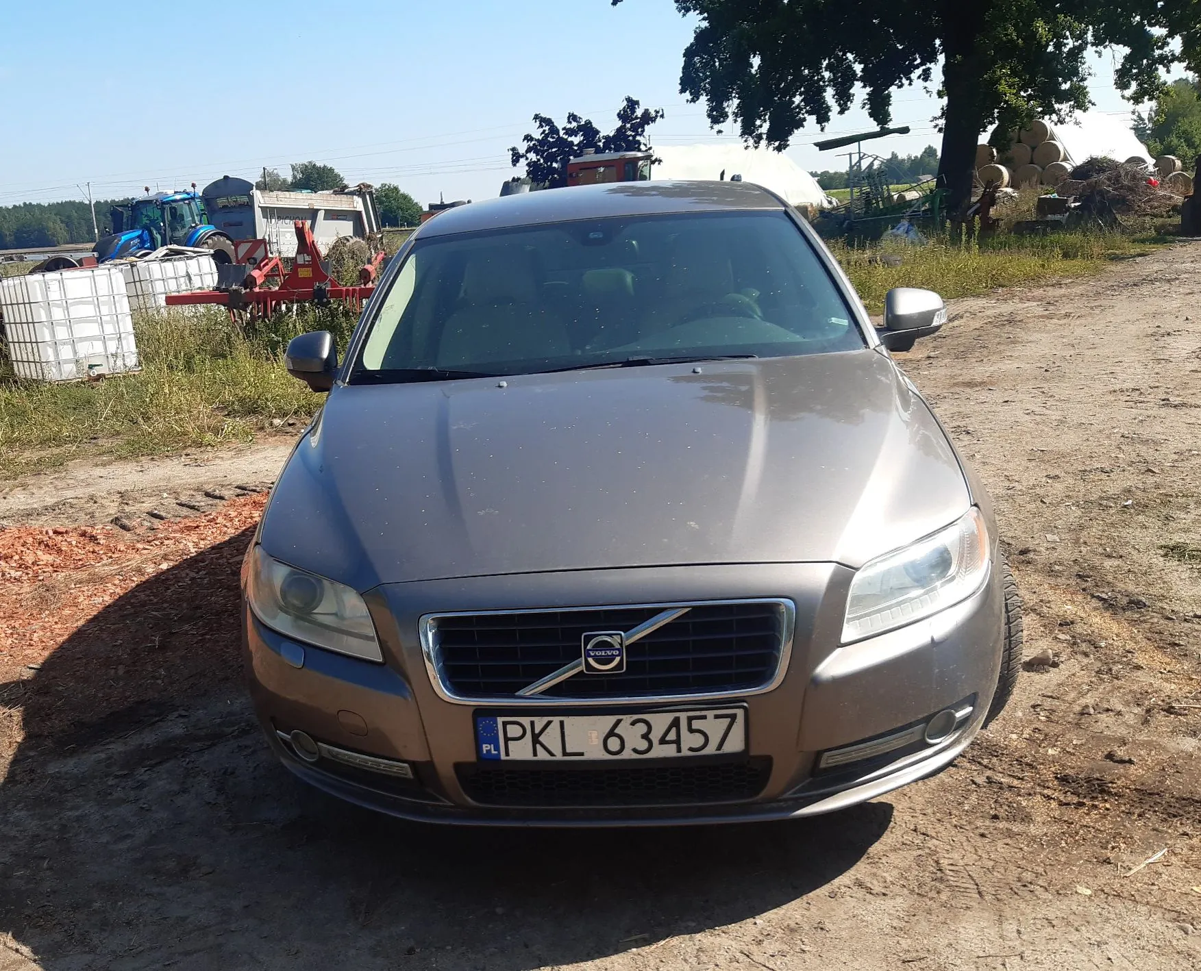 Srebrny samochód marki Volvo zaparkowany na piaszczystej drodze. Na przednim zderzaku znajduje się polska tablica rejestracyjna o numerze PKL 63457. W tle widoczny jest niebieski traktor, białe zbiorniki IBC oraz drzewa pod jasnym, bezchmurnym niebem.