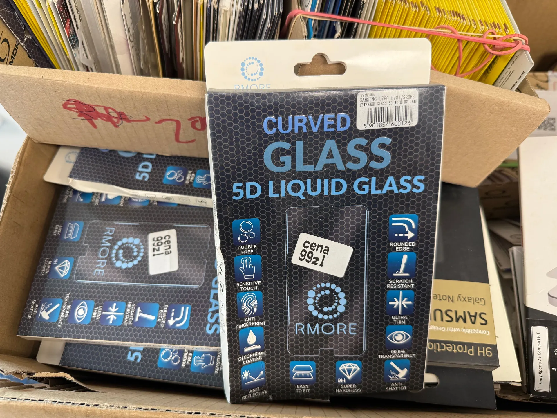 Zdjęcie opakowania ochronnego szkła hartowanego do telefonu marki RMORE. Na opakowaniu widnieje napis 'CURVED GLASS 5D LIQUID GLASS'. Produkt posiada białą etykietę z napisem 'cena 99zł'. Poniżej znajdują się ikony opisujące cechy produktu: 'BUBBLE FREE', 'SENSITIVE TOUCH', 'ANTI FINGERPRINT', 'OLEOPHOBIC COATING', 'ANTI REFLECTIVE', 'EASY TO FIT', 'SUPER HARDNESS', 'ANTI SHATTER', '99.9% TRANSPARENCY', 'ULTRA THIN', 'SCRATCH RESISTANT' oraz 'ROUNDED EDGE'. Opakowanie znajduje się w kartonowym pudełku obok innych podobnych produktów.