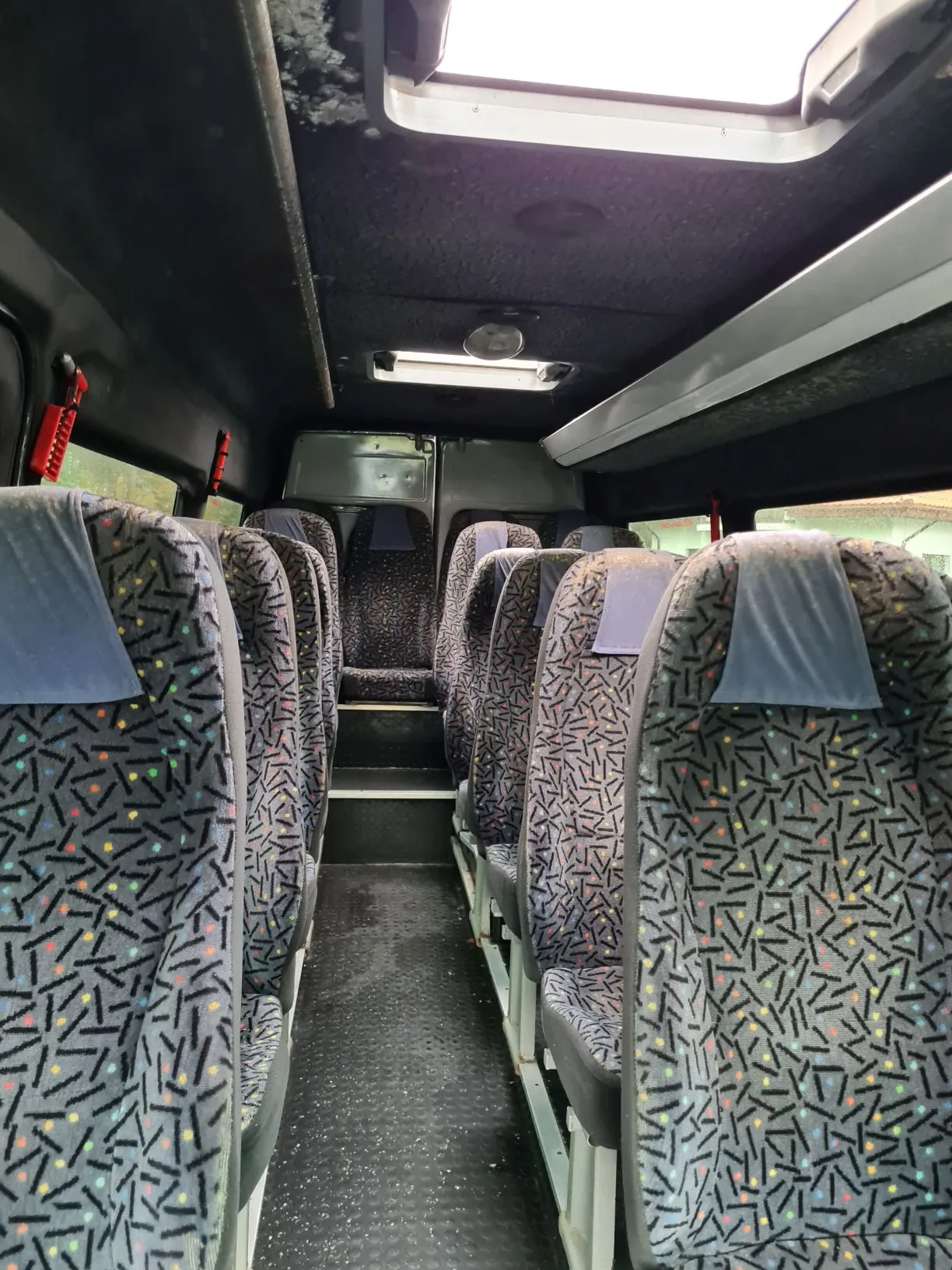 Wnętrze autobusu z rzędami siedzeń o wzorze z czarnymi liniami i kolorowymi kropkami. Siedzenia wyposażone w szare poduszki na oparcia. Po lewej stronie widoczne okno, ciemne ściany i podłoga. Na podłodze znajdują się czerwone przedmioty.