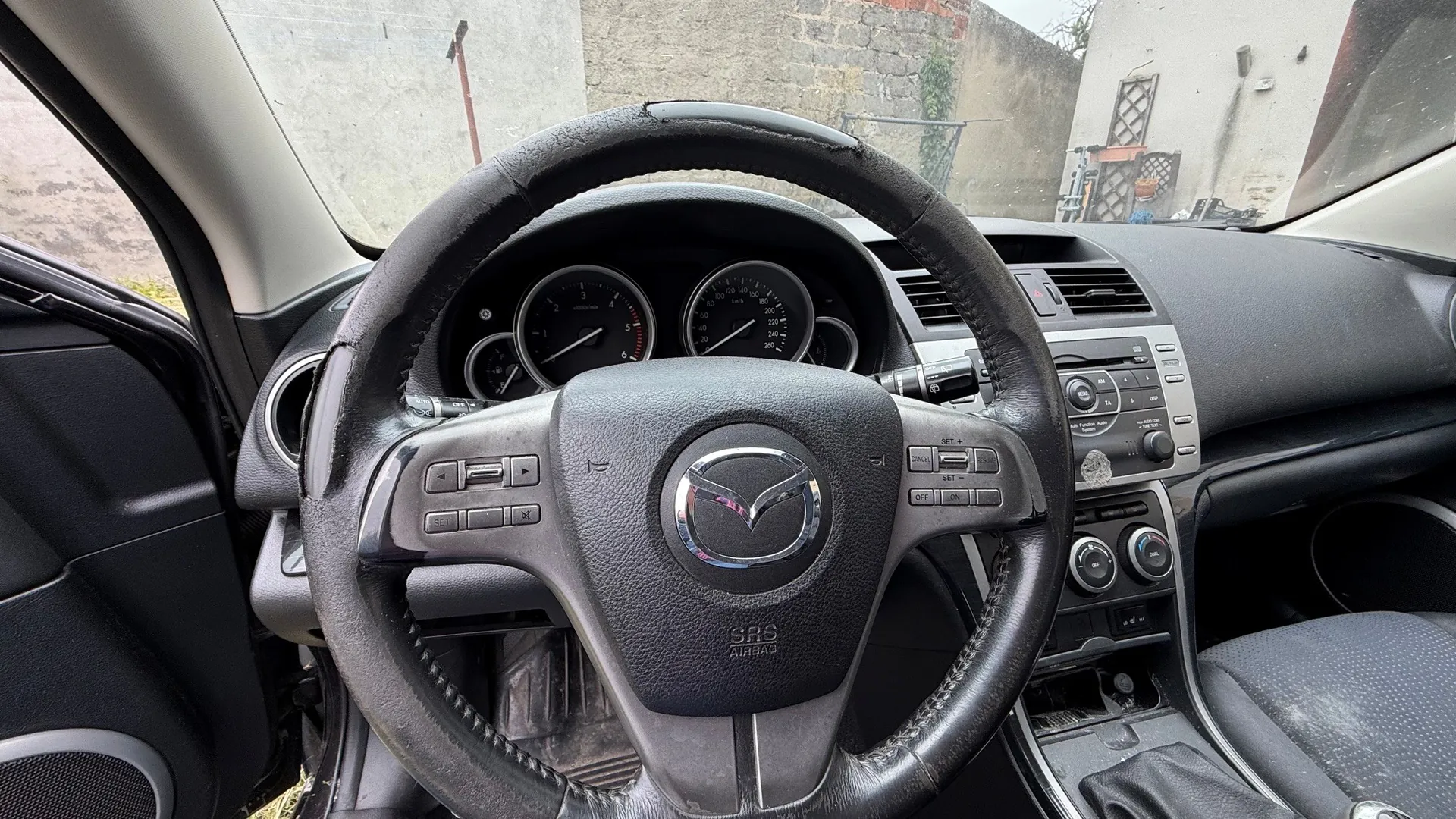 Wnętrze samochodu marki Mazda, widok z perspektywy kierowcy. Na pierwszym planie znajduje się czarna, skórzana kierownica z logo Mazdy na środku i przyciskami sterującymi po bokach. W tle widoczna jest deska rozdzielcza z analogowymi zegarami prędkościomierza i obrotomierza oraz konsola centralna z radiem i pokrętłami klimatyzacji. Przez przednią szybę widoczny jest fragment szarego muru.