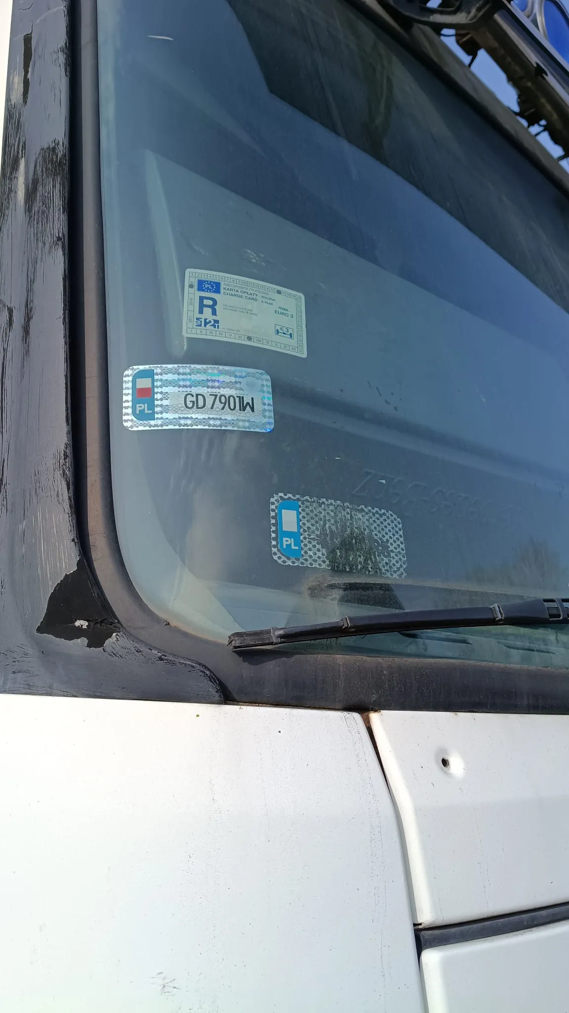 Zbliżenie na przednią szybę białego pojazdu. Na szybie naklejone są trzy elementy: w górnej części biała naklejka z logo i tekstem, w środkowej części polska tablica rejestracyjna o numerze GD 790LW z symbolem PL i flagą Unii Europejskiej, a poniżej mniejsza niebieska naklejka z napisem PL. Widoczna jest również część wycieraczki samochodowej oraz krawędź białej karoserii.