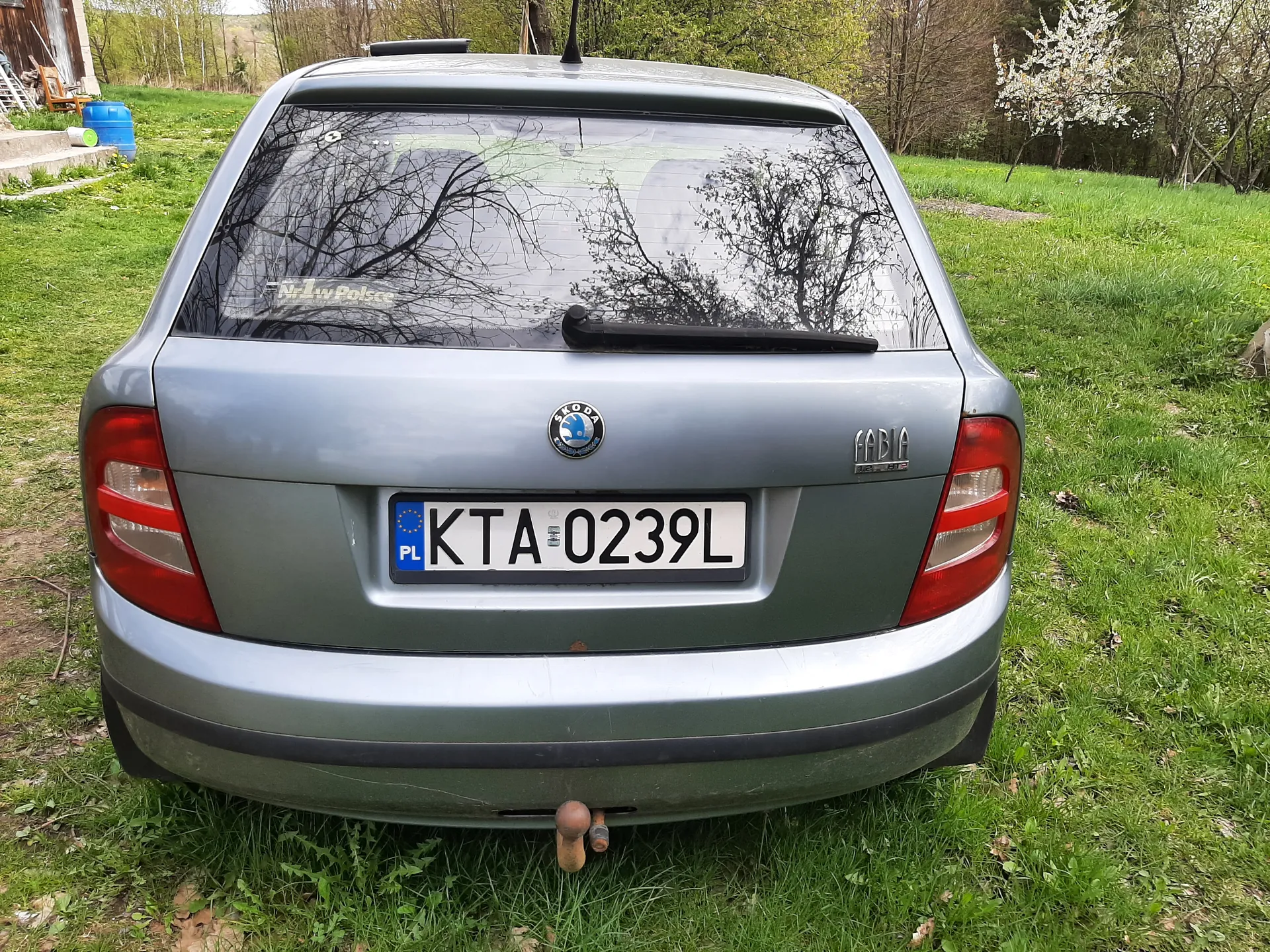 Tył szarego samochodu marki Skoda Fabia zaparkowanego na trawie. Widoczna tablica rejestracyjna z polskim kodem PL i numerem KTA 0239L. Na tylnej szybie odbicie drzew i napisu "Nr1 w Polsce". W tle zielona trawa, drzewa, niebieski beczka oraz fragment budynku. Samochód ma logo Skody i napis "FABIA" po prawej stronie.