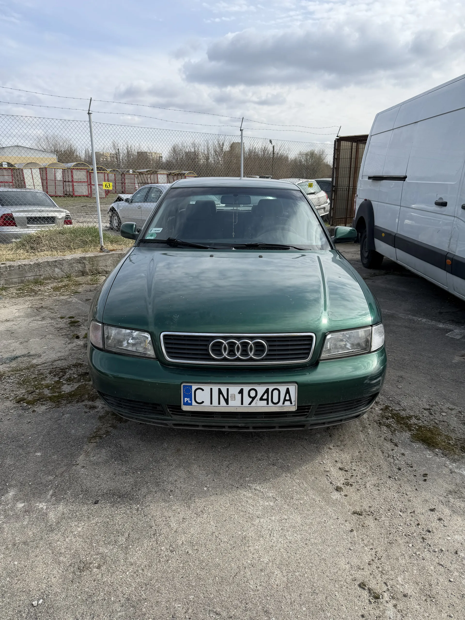 Zielony samochód marki Audi z tablicą rejestracyjną CIN 1940A zaparkowany na podłożu z żwiru. Obok widoczny biały furgon. W tle ogrodzenie siatkowe, inne pojazdy oraz chmurne niebo.