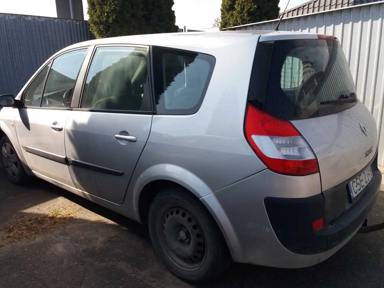 Srebrny samochód osobowy Renault Scenic zaparkowany na podwórku. Widoczna tylna i boczna część pojazdu z czerwonymi światłami tylnymi, tablicą rejestracyjną CS 2748 oraz czarnymi elementami na zderzaku i dolnej części karoserii. W tle metalowe ogrodzenie, drzewa i budynek z dachem. Samochód stoi na nierównym, częściowo zarośniętym podłożu.