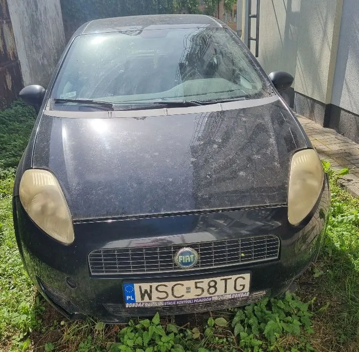 Widok z przodu na czarny samochód marki Fiat. Pojazd jest zakurzony, ma zmatowiałe reflektory i polską tablicę rejestracyjną o numerze WSC 58TG. Samochód stoi na trawie obok budynku.