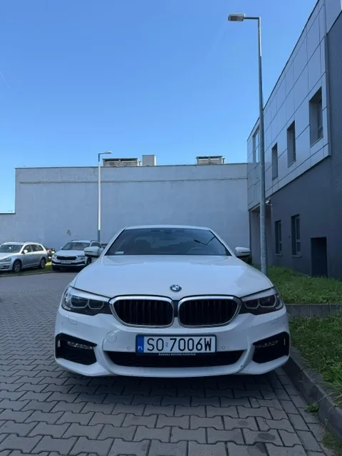 Biały samochód marki BMW zaparkowany przodem do obiektywu na kostce brukowej. Pojazd posiada tablicę rejestracyjną o numerze SO 7006W. W tle widoczny jest szary budynek przemysłowy, latarnia uliczna oraz inne zaparkowane samochody pod bezchmurnym, niebieskim niebem.