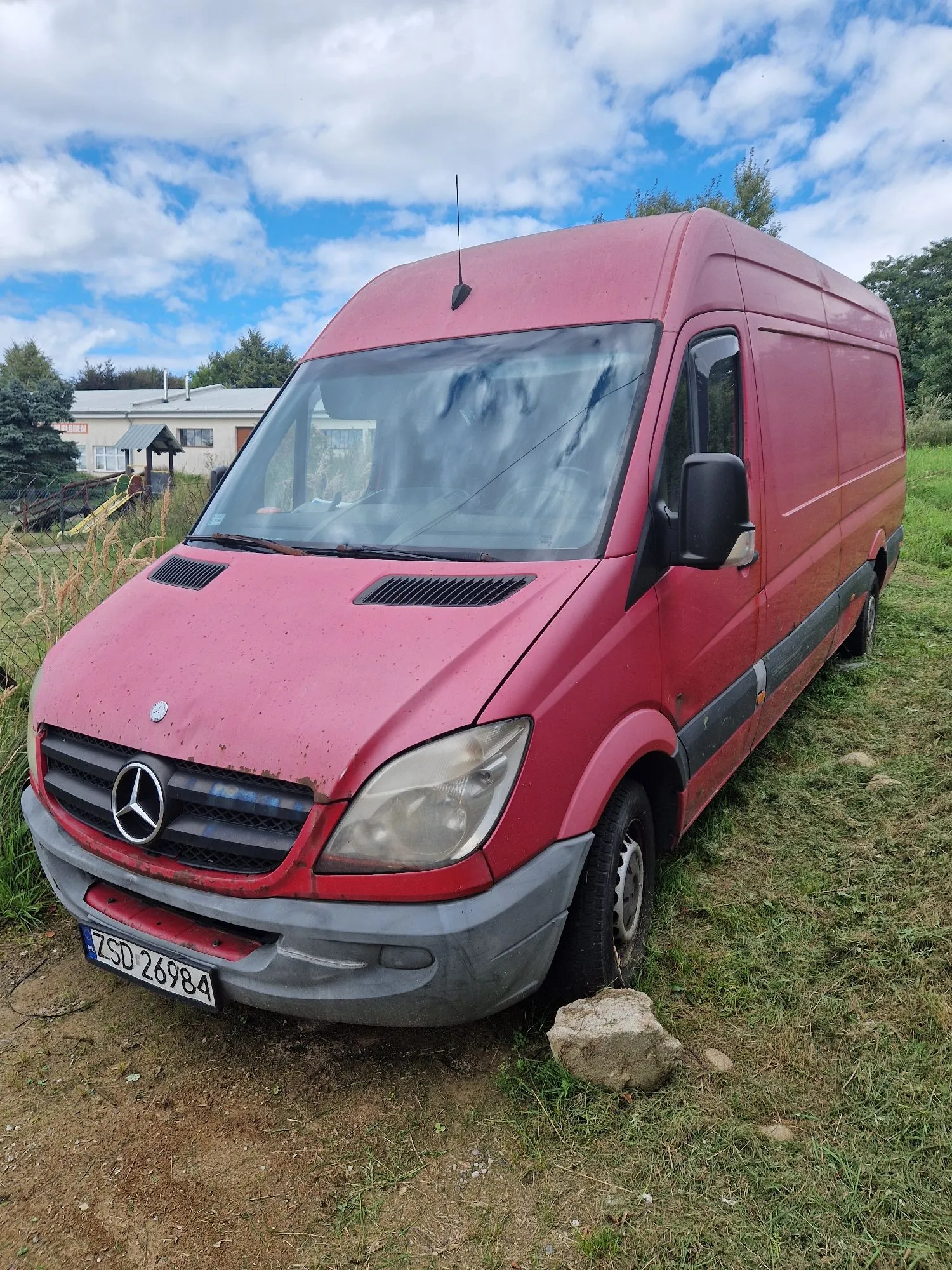 Czerwony furgon marki Mercedes-Benz zaparkowany na trawiastym terenie. Pojazd jest w złym stanie technicznym, z widocznymi śladami zużycia i zabrudzeniami. Na przodzie znajduje się tablica rejestracyjna o numerze ZSD 26984. W tle widać budynek mieszkalny oraz niebo z białymi chmurami.