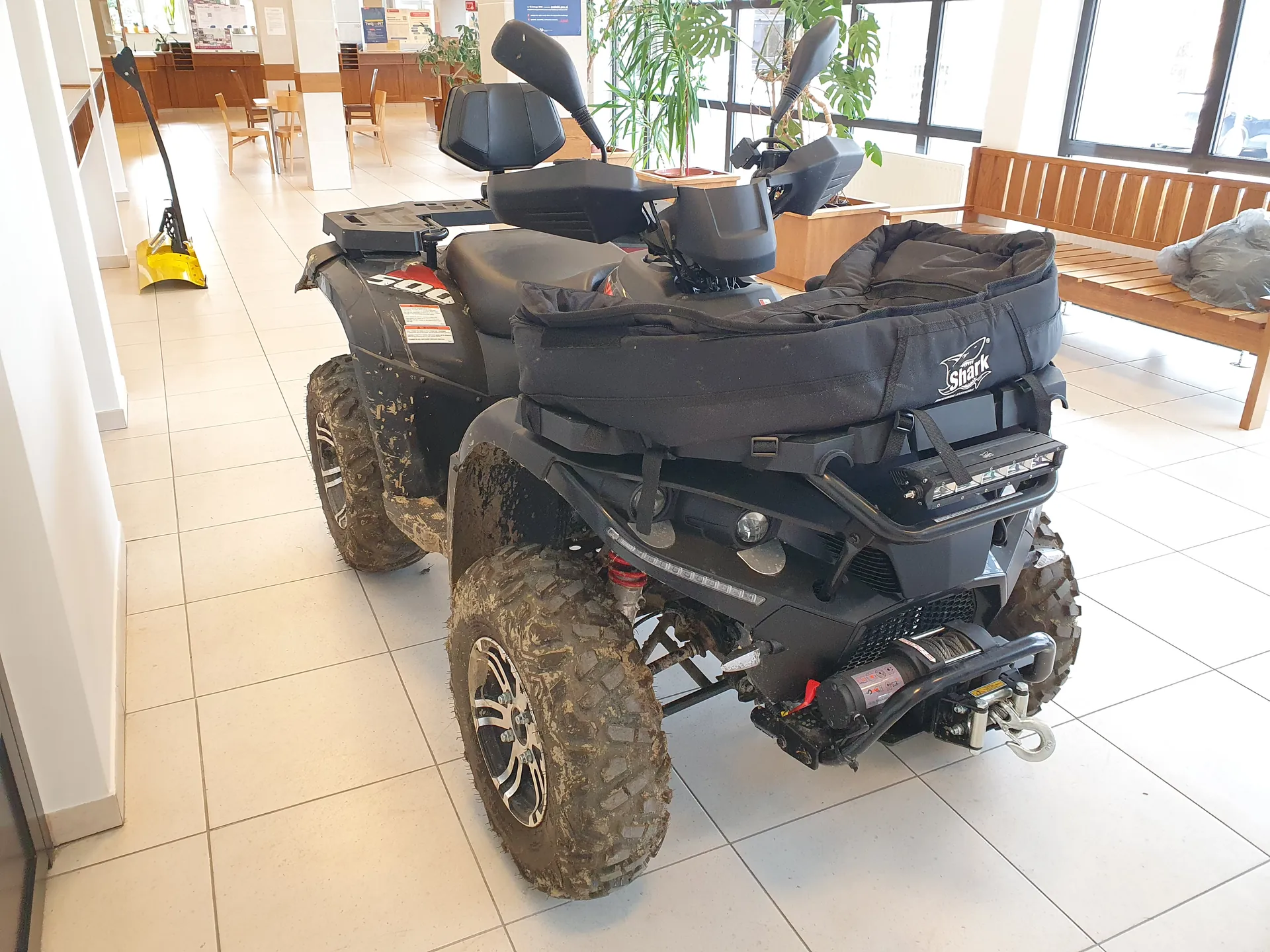 Czarny quad z oznaczeniem 500 i logo Shark na czarnej torbie na bagaż. Quad ma zabrudzone opony, widoczne siedzenia, lusterka i reflektory. Pojazd znajduje się w pomieszczeniu z białą płytkową podłogą, drewnianą ławką, krzesłami, roślinami i oknami. W tle widoczne są tablice informacyjne oraz żółty sprzęt czyszczący.