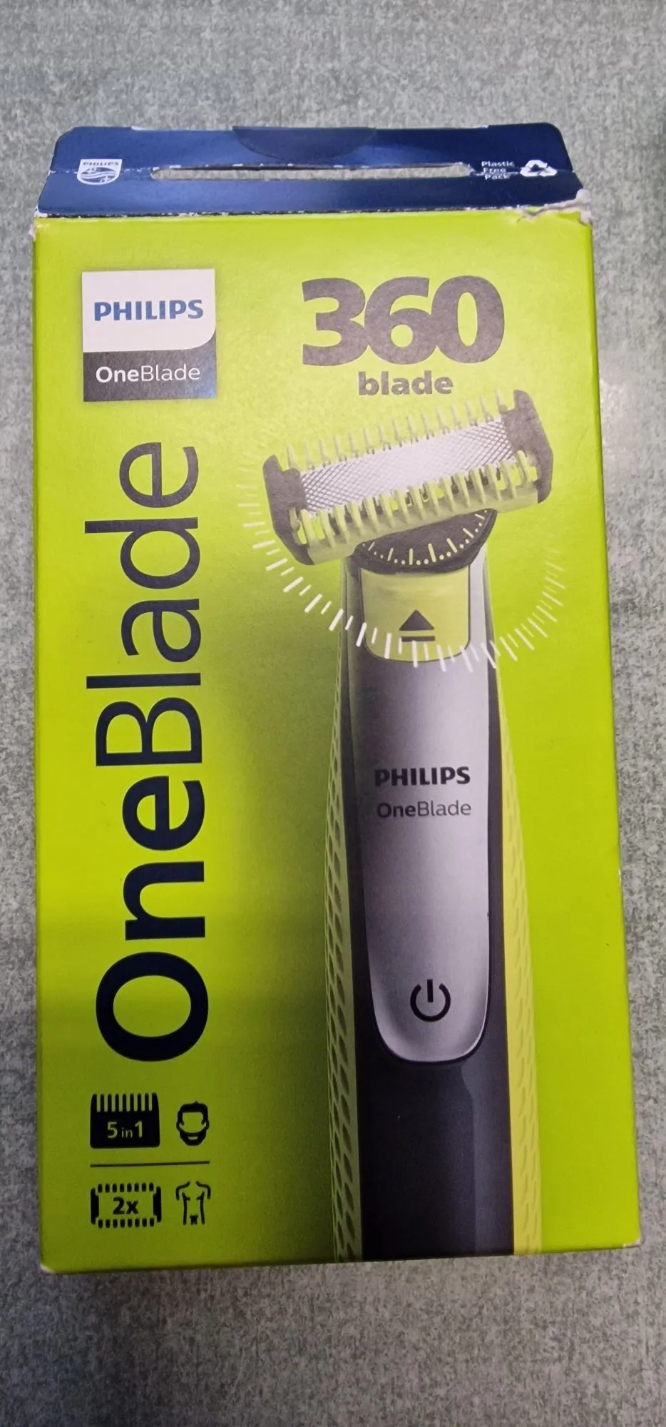 Opakowanie trymera Philips OneBlade w kolorze limonkowym. Na przodzie znajduje się zdjęcie urządzenia oraz duże napisy: 'PHILIPS OneBlade' w pionie po lewej stronie oraz '360 blade' w górnej części. Na dole widoczne są ikony informujące o zawartości zestawu: '5 in 1' oraz '2x' z grafiką ostrzy.