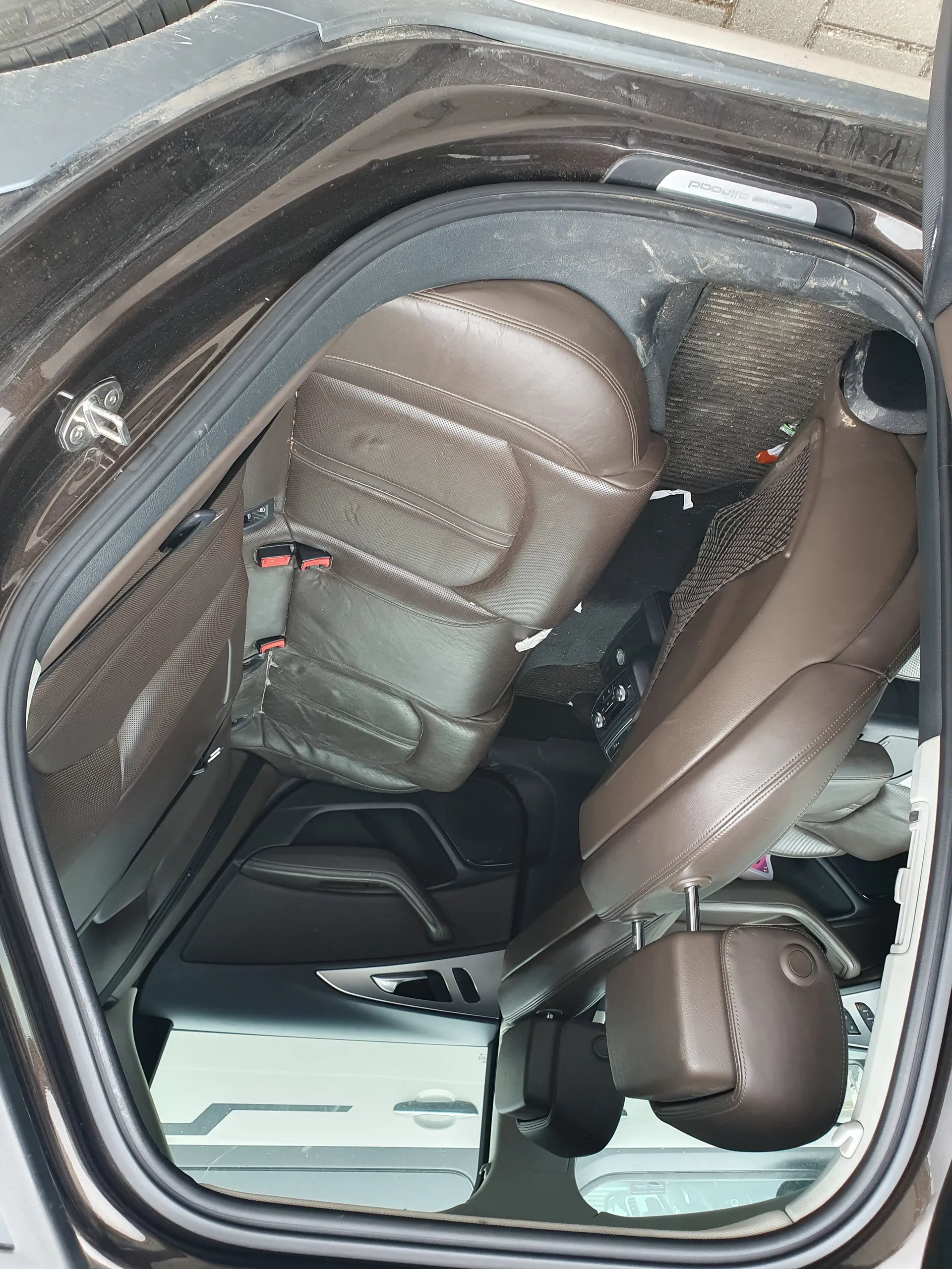 Wnętrze samochodu widziane od strony otwartego tylnego drzwi. Brązowe skórzane siedzenia tylne z czerwonymi zapięciami pasów bezpieczeństwa. Widoczne poduszki pod głowę, brudne dywaniki z resztkami śmieci. Na ramie drzwi widoczny napis "PROTECT". Wnętrze wydaje się nieco zabrudzone, z widocznymi plamami na siedzeniach i drzwiach.