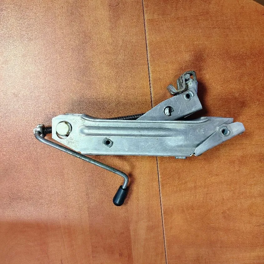 Metalowy mechanizm podnośnika szyby samochodowej z korbą, leżący na brązowej, drewnianej powierzchni.