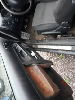 Wnętrze samochodu z otwartą drzwiami kierowcy. Widoczna deska rozdzielcza, kierownica po prawej stronie, czarne siedzenie oraz podłoga z widocznymi śladami brudu. Drzwi posiadają drewnianą obudowę z widocznymi uszkodzeniami. Poza samochodem widoczny jest żwirowy podjazd.