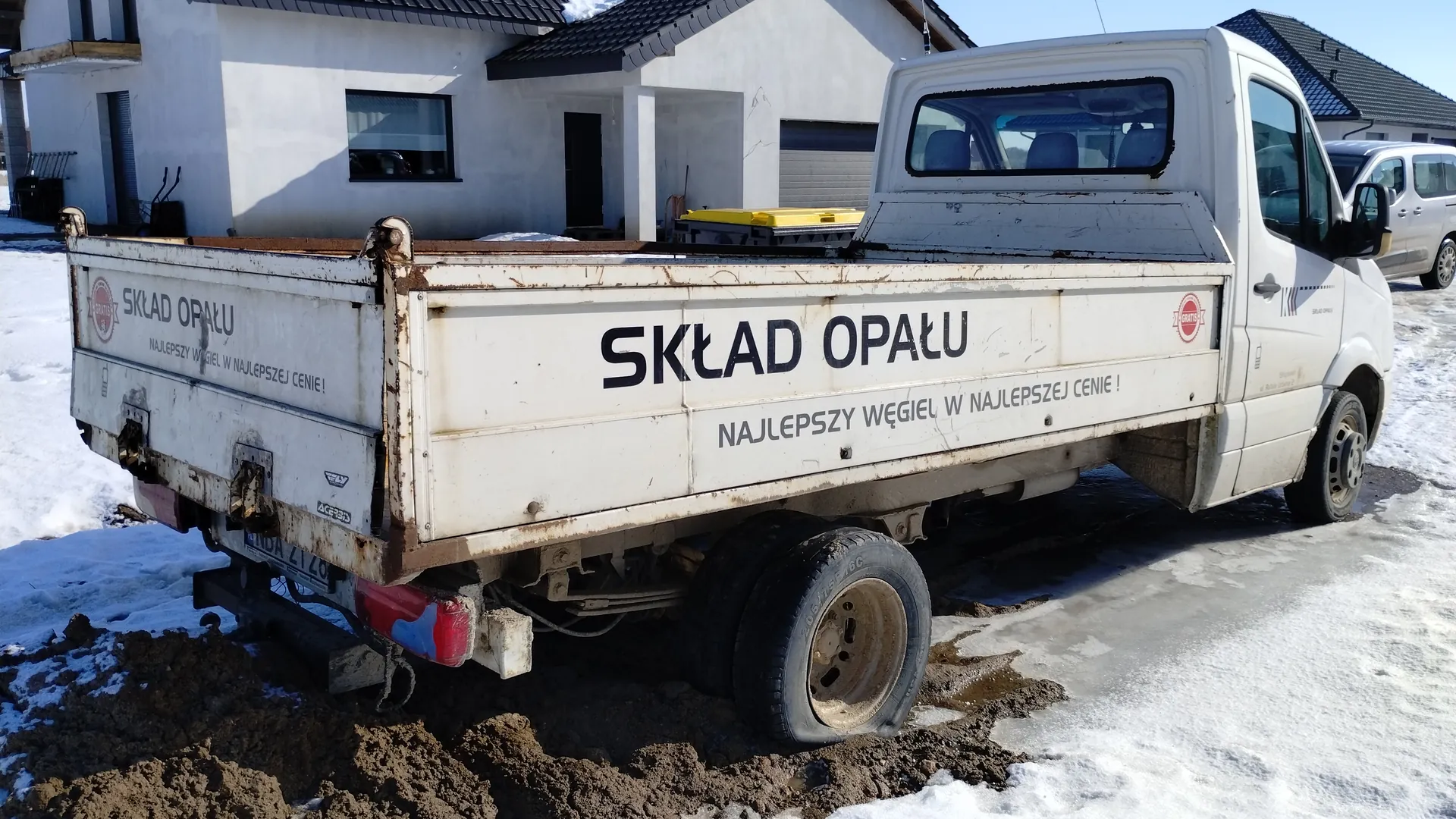 Biały samochód dostawczy z napisem "SKŁAD OPAŁU" oraz "NAJLEPSZY WĘGIEL W NAJLEPSZEJ CENIE!" na boku i tylnej ścianie. Pojazd stoi na zasypanej śniegiem i błotem powierzchni. W tle widoczne są białe domy z ciemnymi dachami oraz częściowo widoczny srebrny samochód. Na tylnym zderzaku znajduje się tablica rejestracyjna, a na dnie ładowni leży żółty pojemnik. Samochód ma ślady zużycia i rdzy.