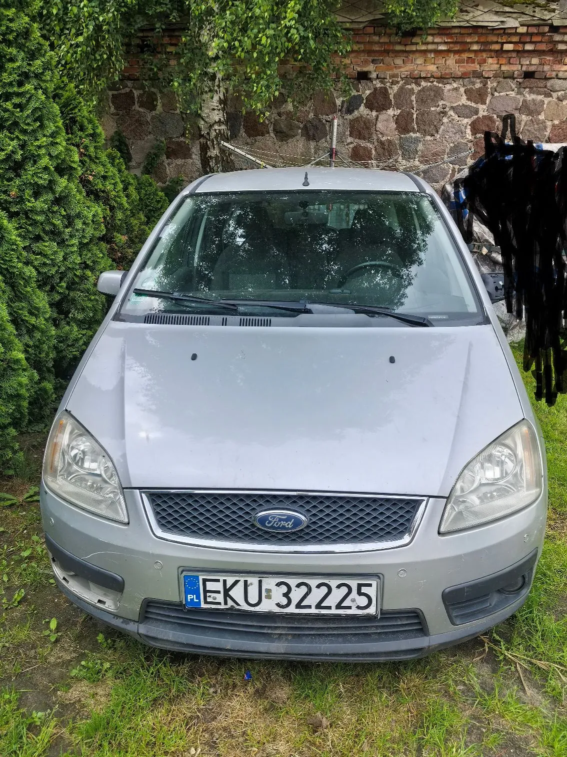 Srebrny samochód marki Ford widziany od przodu, zaparkowany na trawie przed kamiennym murem i zielonymi krzewami. Pojazd posiada polską tablicę rejestracyjną o numerze EKU 32225.
