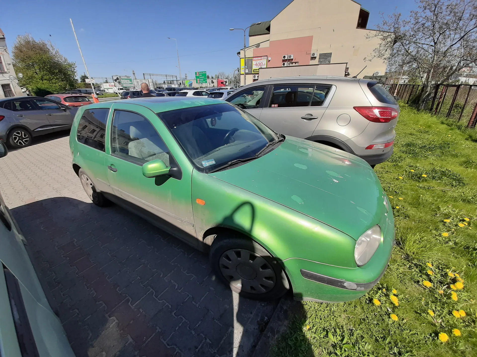 Zielony samochód osobowy zaparkowany na brukowanym placu, obok srebrnego SUV-a. W tle inne zaparkowane pojazdy, budynki, w tym budynek z żółtym znakiem z napisem "400" oraz zielonymi tablicami drogowymi. Po prawej stronie trawa z żółtymi kwiatami (mniszkiem), jasne niebo. Na pierwszym planie cień od obiektu poza kadrem.
