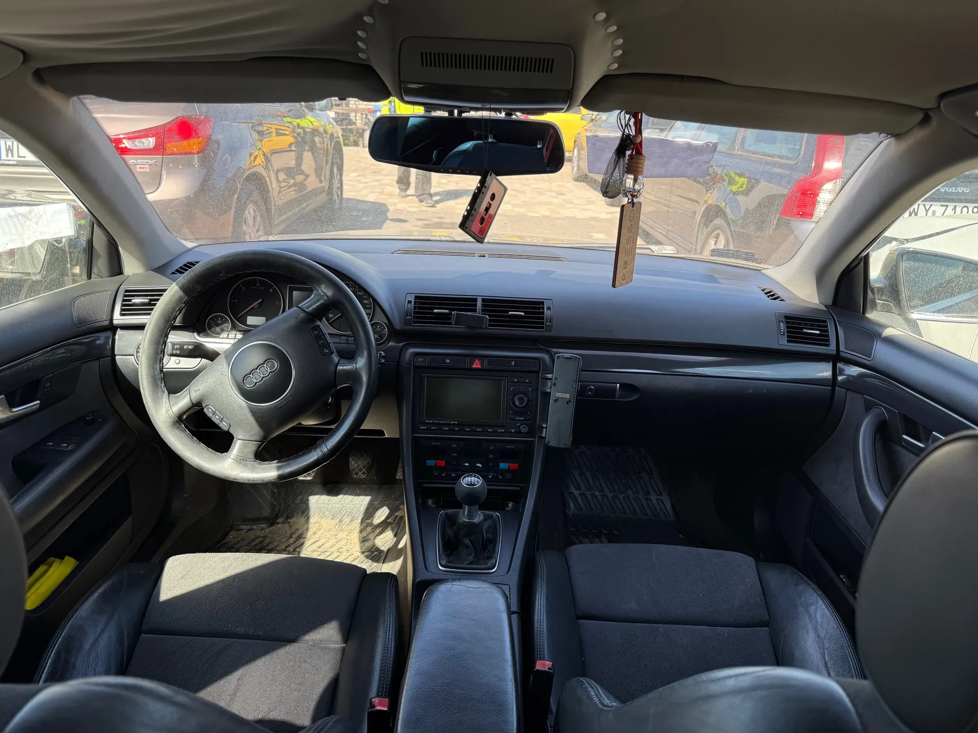 Wnętrze samochodu marki Audi z widoczną kierownicą z logo, deską rozdzielczą z licznikami, centralną konsolą z ekranem i przyciskami, dźwignią zmiany biegów oraz czarnymi skórzonymi fotelami. W lusterku wstecznym wiszą przedmioty: kaseta magnetofonowa i drewniana tabliczka. Przez przednią szybę widoczne są zaparkowane samochody, w tym niebieski SUV i żółty pojazd.