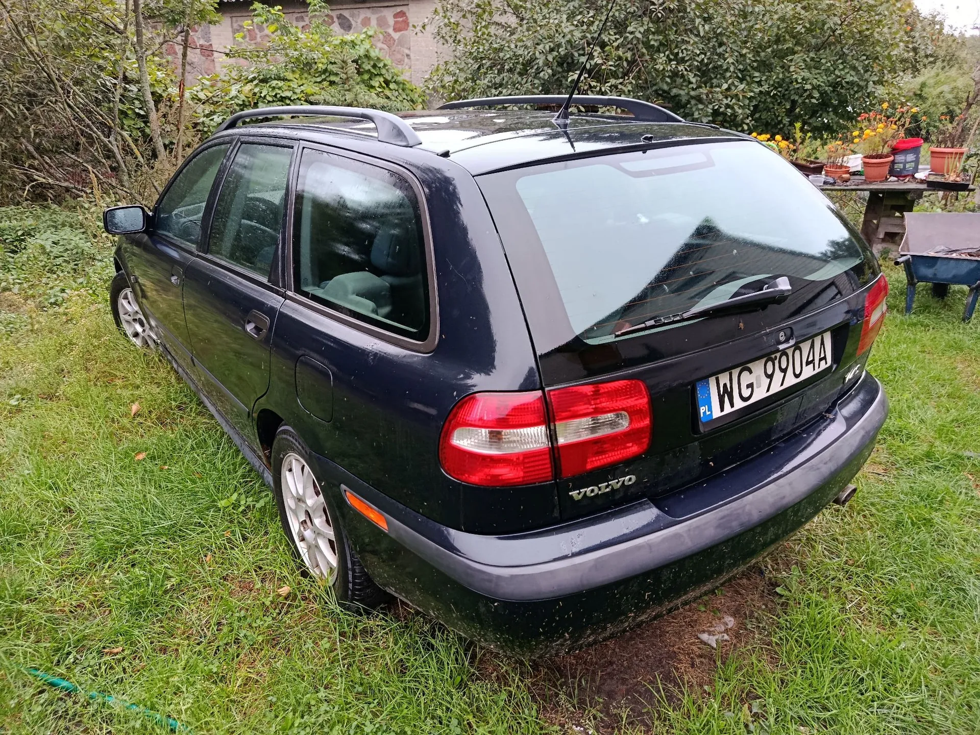 Ciemnogranatowe Volvo kombi zaparkowane na trawiastym terenie. Widoczny tył i lewy bok samochodu. Auto posiada relingi dachowe oraz polską tablicę rejestracyjną o numerze WG 9904A. W tle znajdują się drzewa, krzewy oraz doniczki z roślinami.