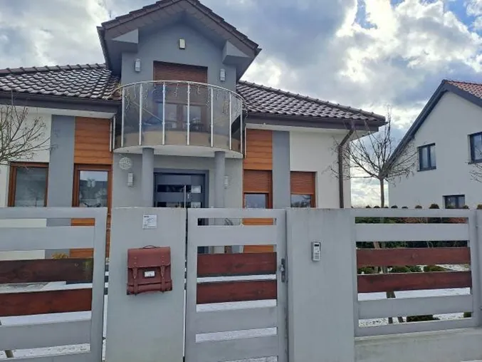 Nowoczesny, parterowy dom jednorodzinny z poddaszem, wykończony w kolorach szarym, białym i z elementami drewnianymi na elewacji. Dom posiada balkon z metalową balustradą oraz ciemnobrązowy dach z dachówek. Przed domem znajduje się szary ogrodzenie z poziomymi, drewnianymi wstawkami oraz brama. Na ogrodzeniu zamontowana jest brązowa skrzynka pocztowa i domofon. W tle widoczny jest fragment innego białego domu oraz zachmurzone niebo.
