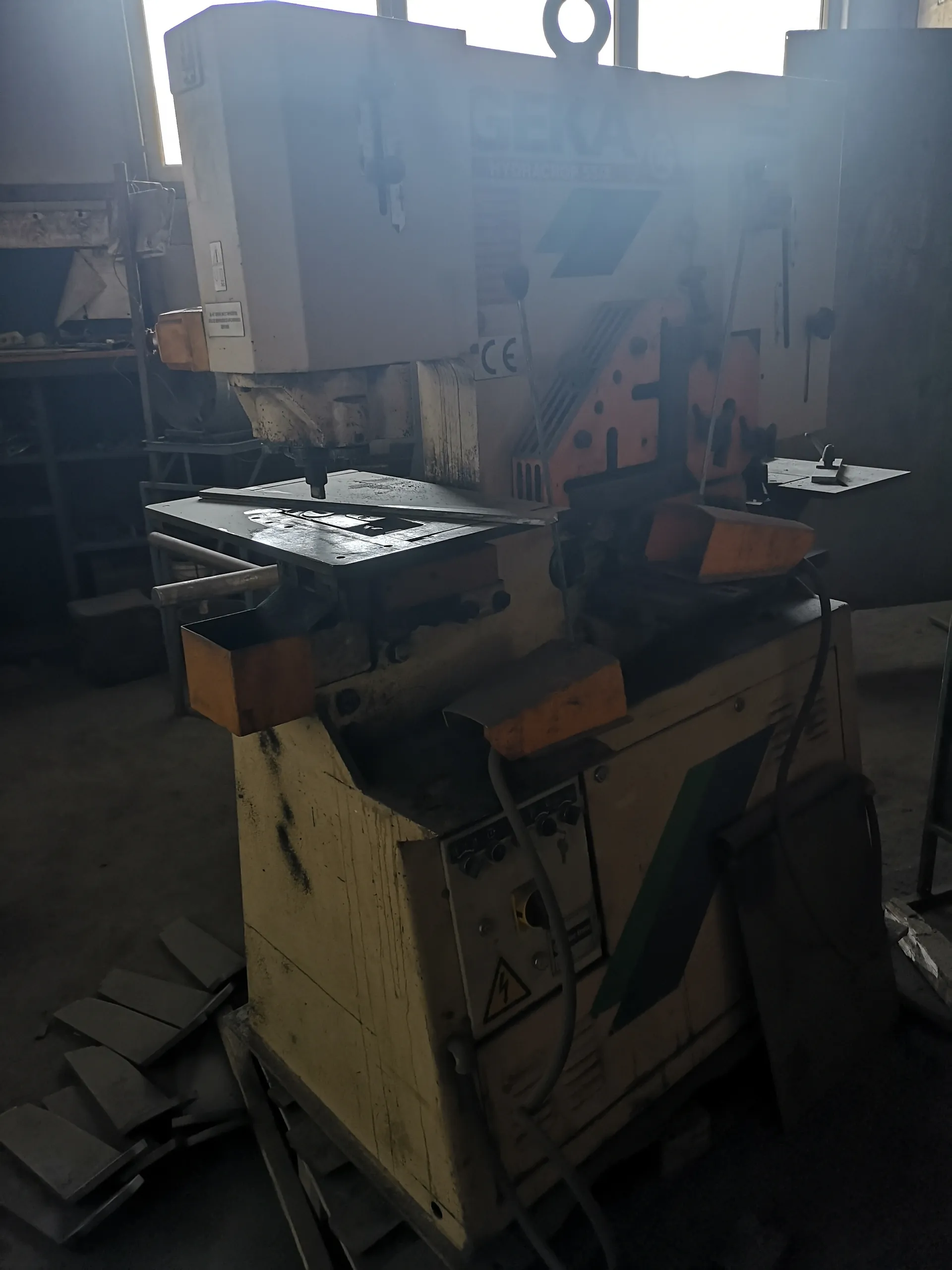 Przemysłowa maszyna cięcia hydrauliczna marki GEKA model HYDRA-CROP 55/A z widocznym znakiem CE. Maszyna ma zniszczoną, zardzewiałą powierzchnię, panel sterowania z przyciskami i ostrzeżeniem przed napięciem. W tle warsztat z półkami, narzędziami i metalowymi elementami na podłodze. Na maszynie widoczne są pomarańczowe i zielone elementy oraz przewody.