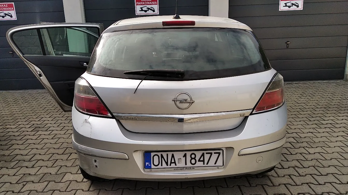 Srebrny samochód marki Opel Astra widziany od tyłu, zaparkowany na kostce brukowej przed szarymi bramami garażowymi. Lewe tylne drzwi są otwarte. Na klapie bagażnika znajduje się logo Opela oraz napis 'ASTRA' i '1.6'. Tablica rejestracyjna ma numer 'ONA 18477' z oznaczeniem 'PL'. Nad garażami widoczne są znaki zakazu parkowania.