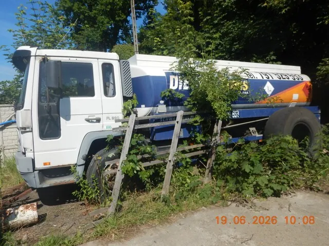 Biała ciężarówka z dużym, niebiesko-białym zbiornikiem na płynach, zaparkowana w zarośniętym terenie. Zbiornik posiada napis 'D...' oraz numer telefonu '791 525 333'. Pojazd jest częściowo zasłonięty przez drewniany płot i gęste zielone krzewy. W prawym dolnym rogu zdjęcia widnieje czerwona data i godzina: '19.06.2026 10:08'.