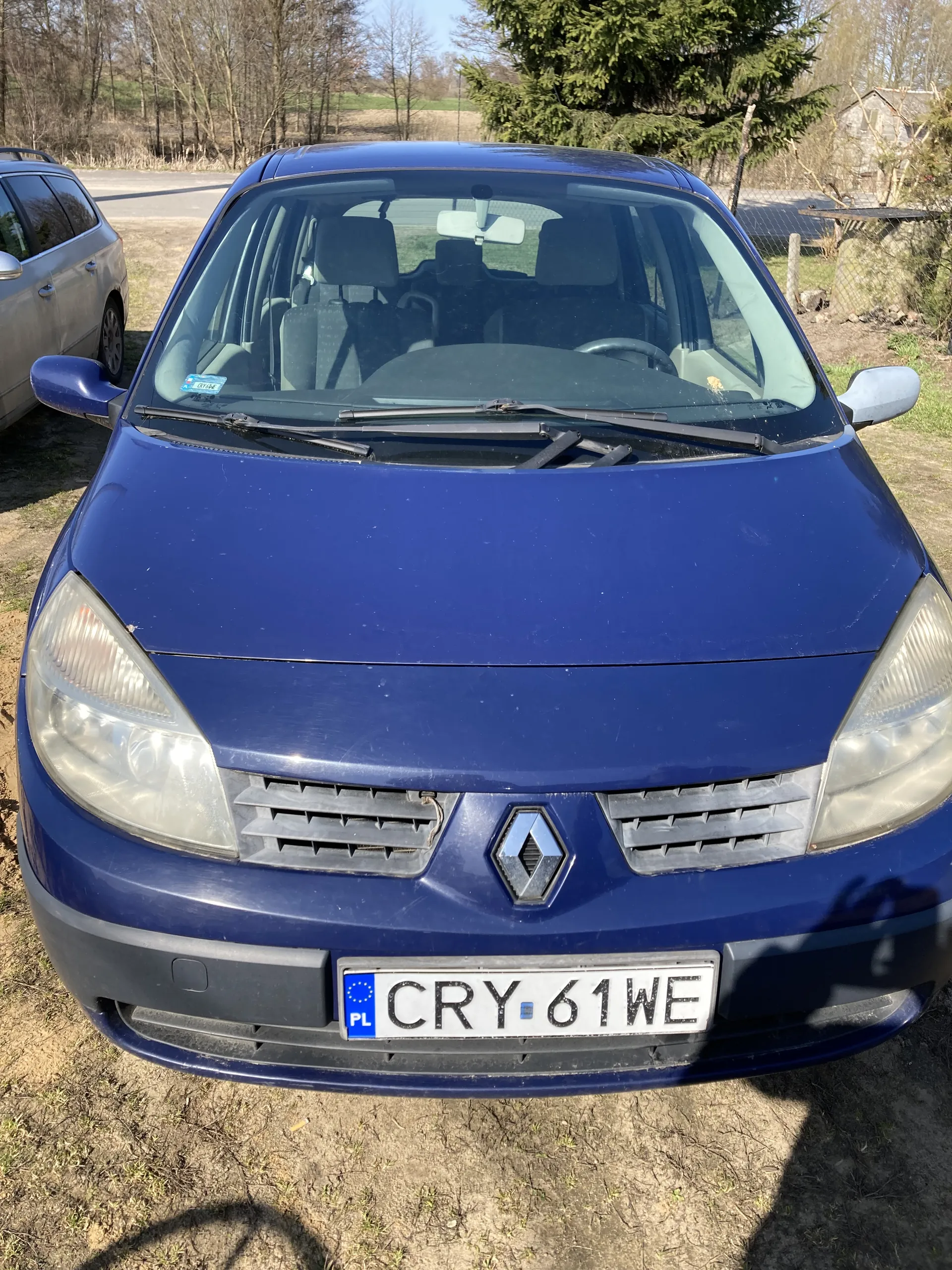 Niebieski samochód marki Renault z przodu, z widoczną tablicą rejestracyjną CRY 61WE oraz oznaczeniem PL. W tle drugi samochód, drzewa, ogrodzenie i dom. Samochód stoi na trawiastym terenie.
