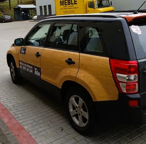Czarny samochód typu SUV zaparkowany na kostce brukowej. Boki pojazdu są oklejone folią imitującą jasnobrązowe drewno. Na drzwiach kierowcy widnieje napis: 'MEBLE NA WYMIAR tel. 509 144 244' oraz logo firmy. W tle widoczny jest żółty samochód dostawczy z napisem 'MEBLE' oraz budynek firmy.