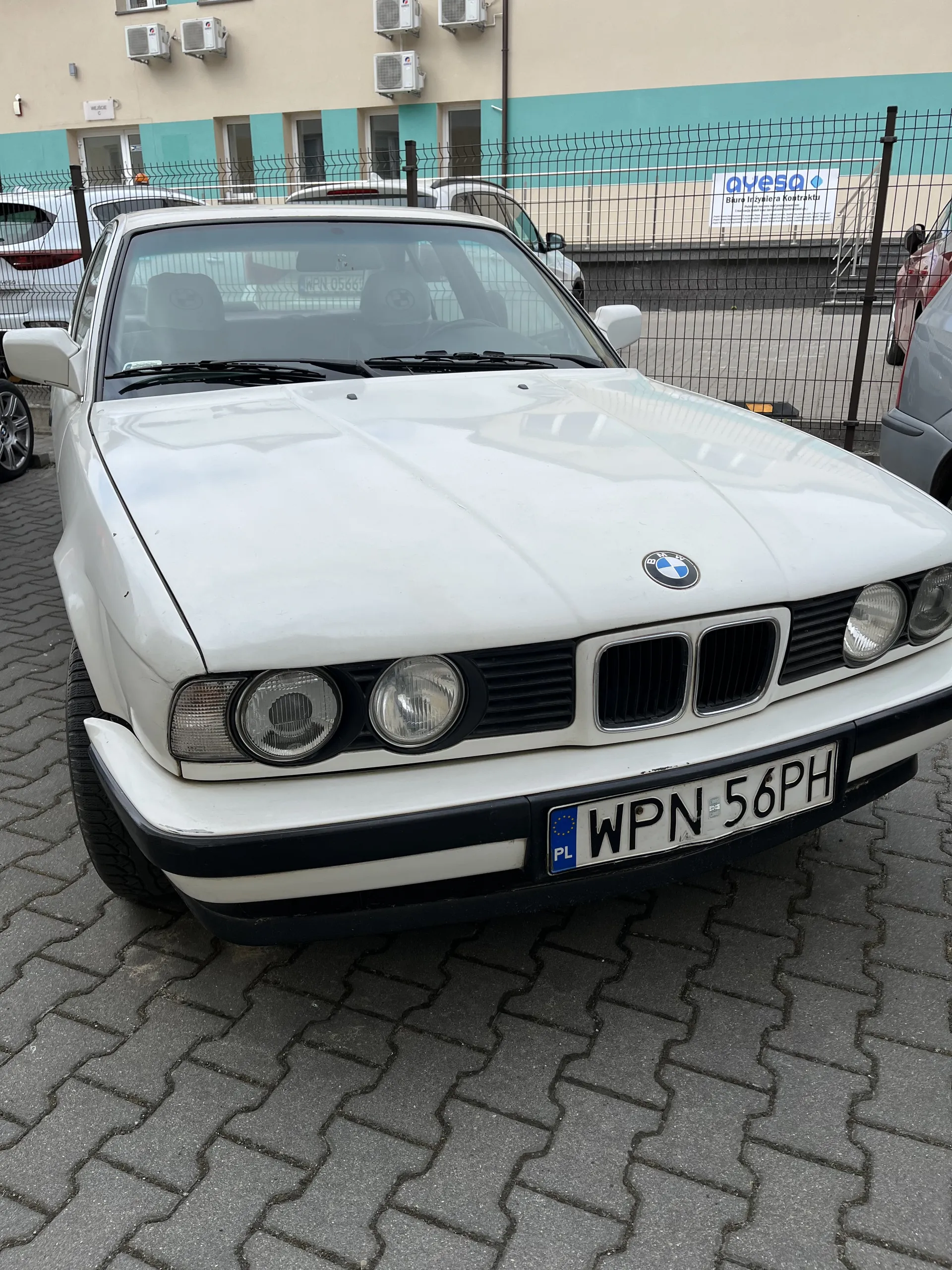 Biały samochód marki BMW z tablicą rejestracyjną WPN 56PH (z niebieskim pasem UE i oznaczeniem PL) zaparkowany na brukowanej powierzchni. Widoczne są reflektory, logo BMW na masce oraz przednia szyba. W tle budynek z turkusowo-beżową elewacją, klimatyzatory, metalowe ogrodzenie oraz tablica z napisem "avesa".