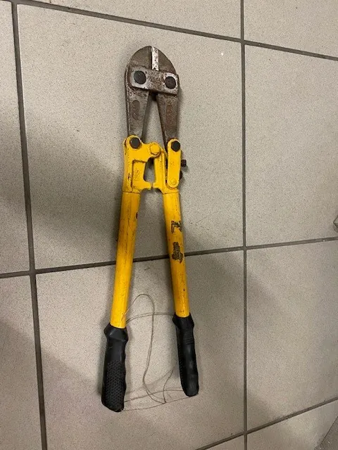 Zardzewiałe nożyce do cięcia prętów z żółtymi uchwytami i czarnymi gumowanymi rękojeściami, umieszczone pionowo na tle jasnoszarej płytkowej ściany z ciemnymi szwami.