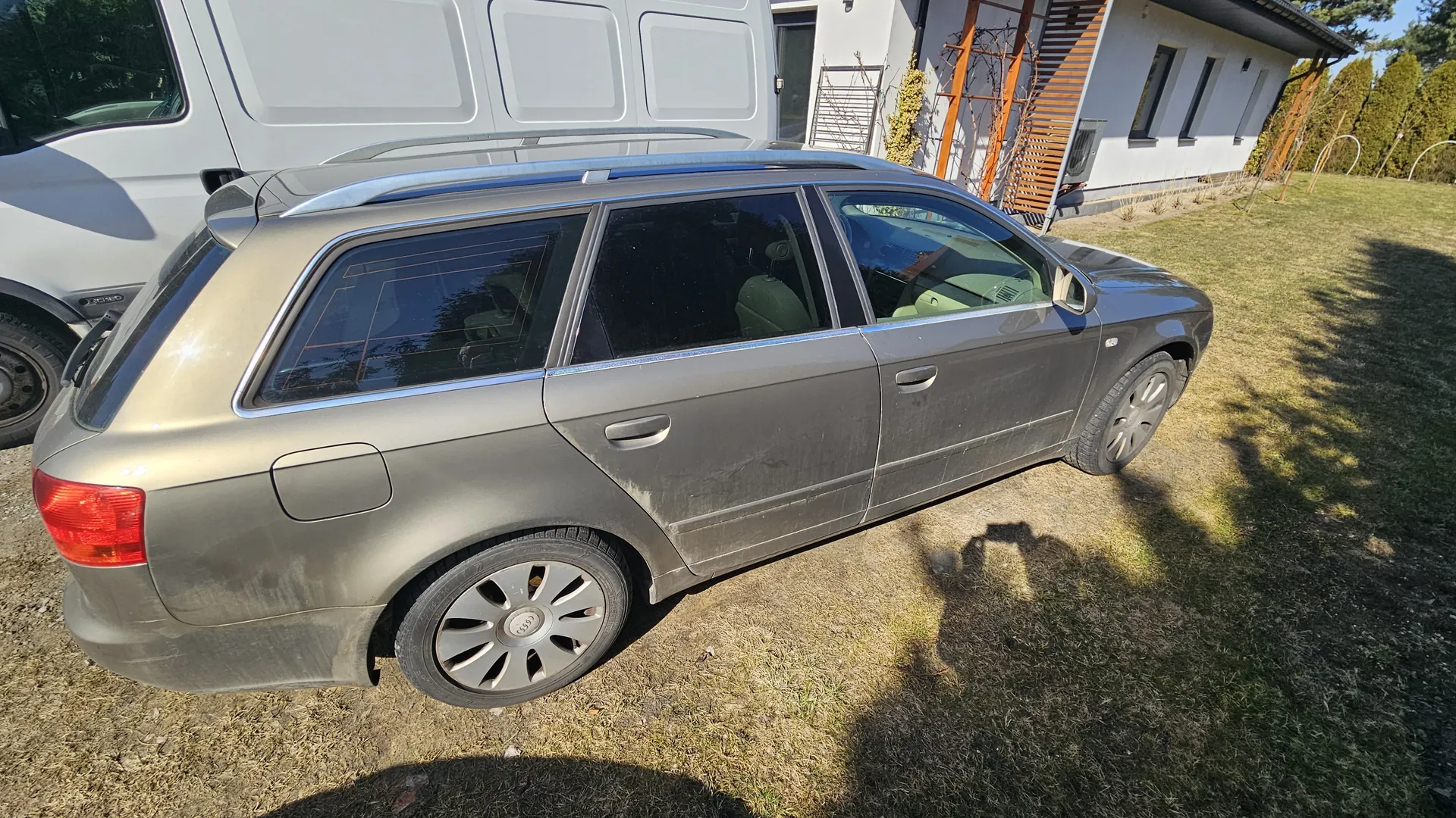 Szary samochód Audi typu kombi z listwami dachowymi, zaparkowany na trawie i żwirze. Po lewej stronie widoczny biały van, w tle biały dom z drewnianą konstrukcją oraz drzewa. Samochód jest zabrudzony, a na kołach widoczne logo Audi. Trawa jest częściowo wysuszona, a na ziemi widoczne cienie.