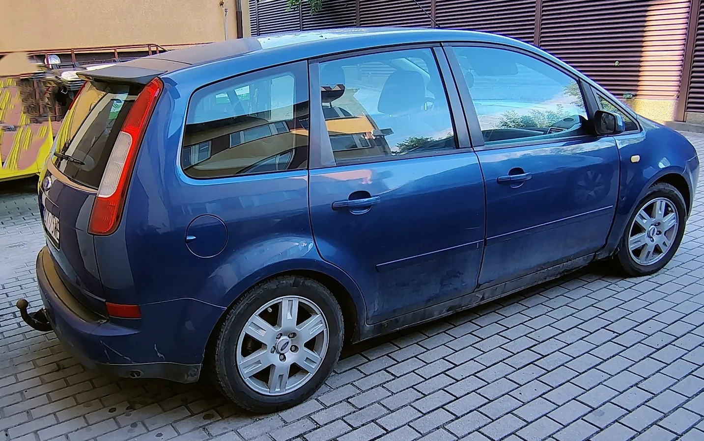 Niebieski samochód osobowy typu hatchback zaparkowany na kostce brukowej. Pojazd jest pokazany z perspektywy trzech czwartych od tyłu, z boku widać drzwi pasażera i tylne drzwi. W tle znajduje się beżowa ściana budynku oraz ciemnobrązowa roleta sklepowa.