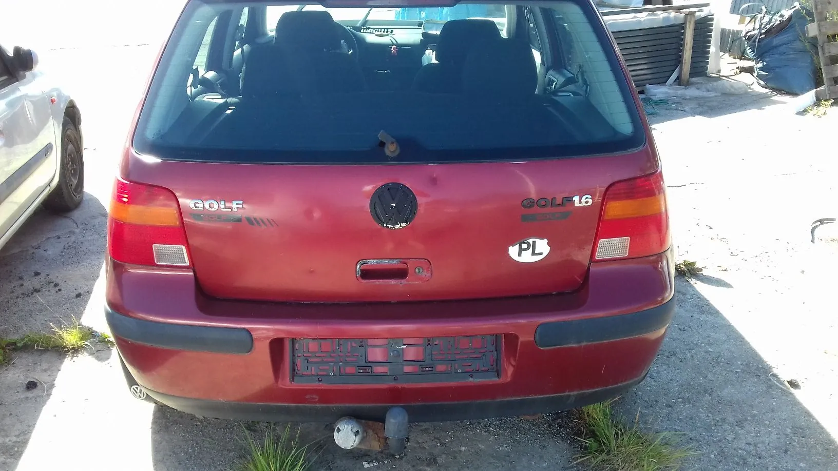 Tylny widok czerwonego samochodu marki Volkswagen Golf. Na klapie bagażnika widoczne są napisy 'GOLF' po lewej stronie oraz 'GOLF 16' po prawej stronie. Po prawej stronie znajduje się również owalna naklejka z literami 'PL'. Samochód posiada zamontowany hak holowniczy. W tle widoczny jest fragment innego białego samochodu oraz betonowe podłoże.