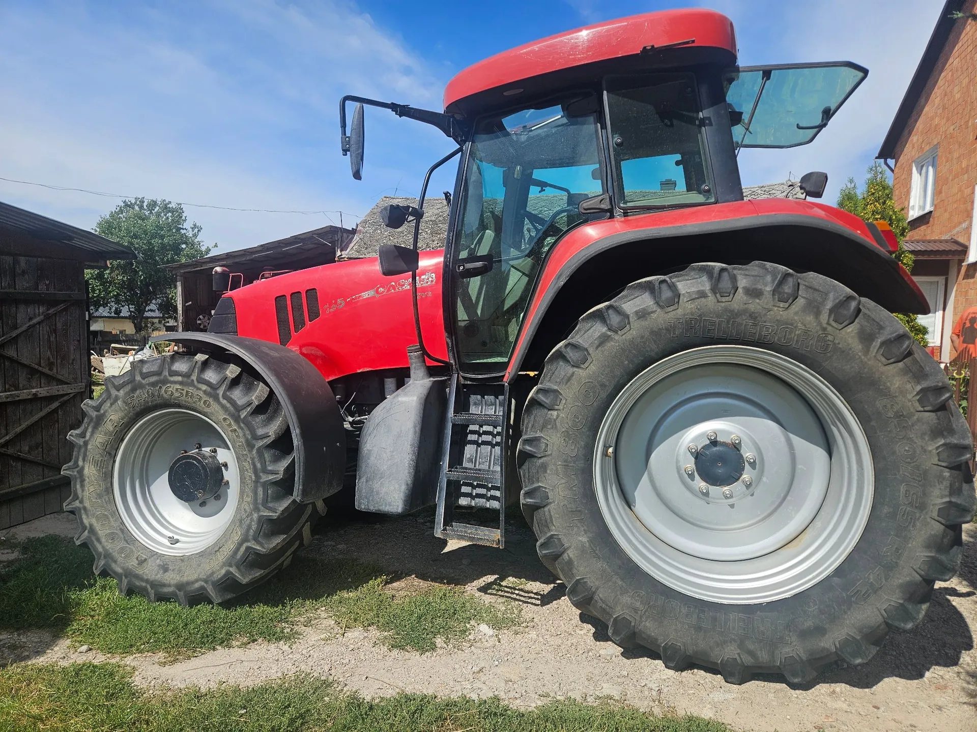 Czerwony traktor rolniczy zaparkowany na trawiastym podłożu przed budynkami. Pojazd posiada duże, czarne opony z wyraźnym bieżnikiem i srebrnymi felgami. Widoczna jest przeszklona kabina kierowcy oraz schodki prowadzące do wejścia.