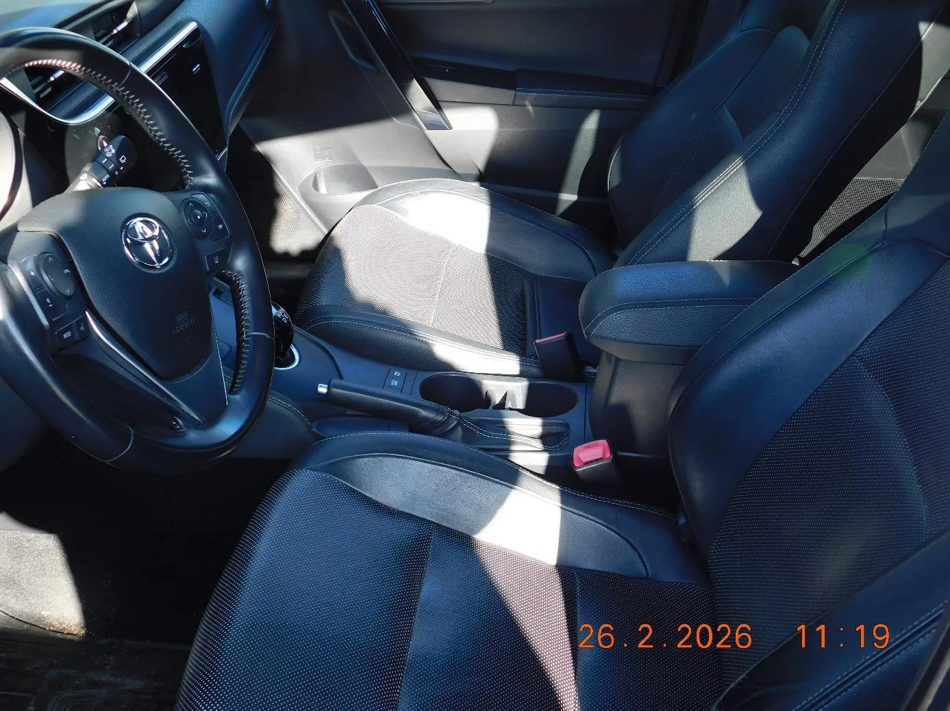 Wnętrze samochodu marki Toyota z czarnymi fotelami, kierownicą z logo Toyoty, centralną konsolą z dźwignią zmiany biegów i uchwytem na kubki. Widoczny znaczek z datą i godziną: 26.2.2026 11:19