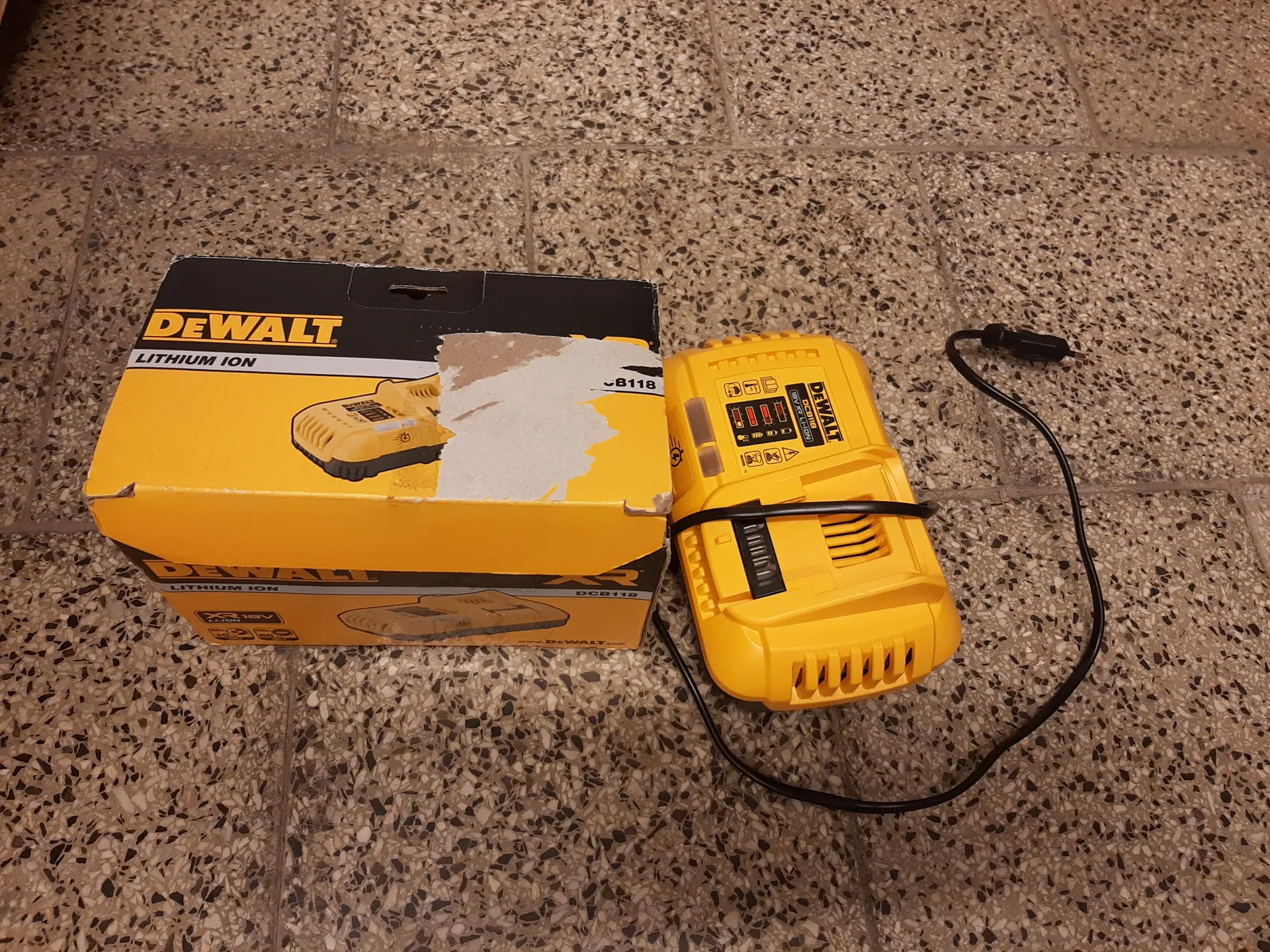 Na podłodze z kafelkami w kolorze beżowym z czarnymi i białymi kropkami leży żółte pudełko marki DEWALT z napisem LITHIUM ION oraz uszkodzonym fragmentem opakowania. Obok pudełka znajduje się żółty ładowarka DEWALT z czarnym kablem z wtyczką. Na ładowarce widoczne są symbole i wskaźniki stanu ładowania. Na opakowaniu widoczny jest również model DCB118.